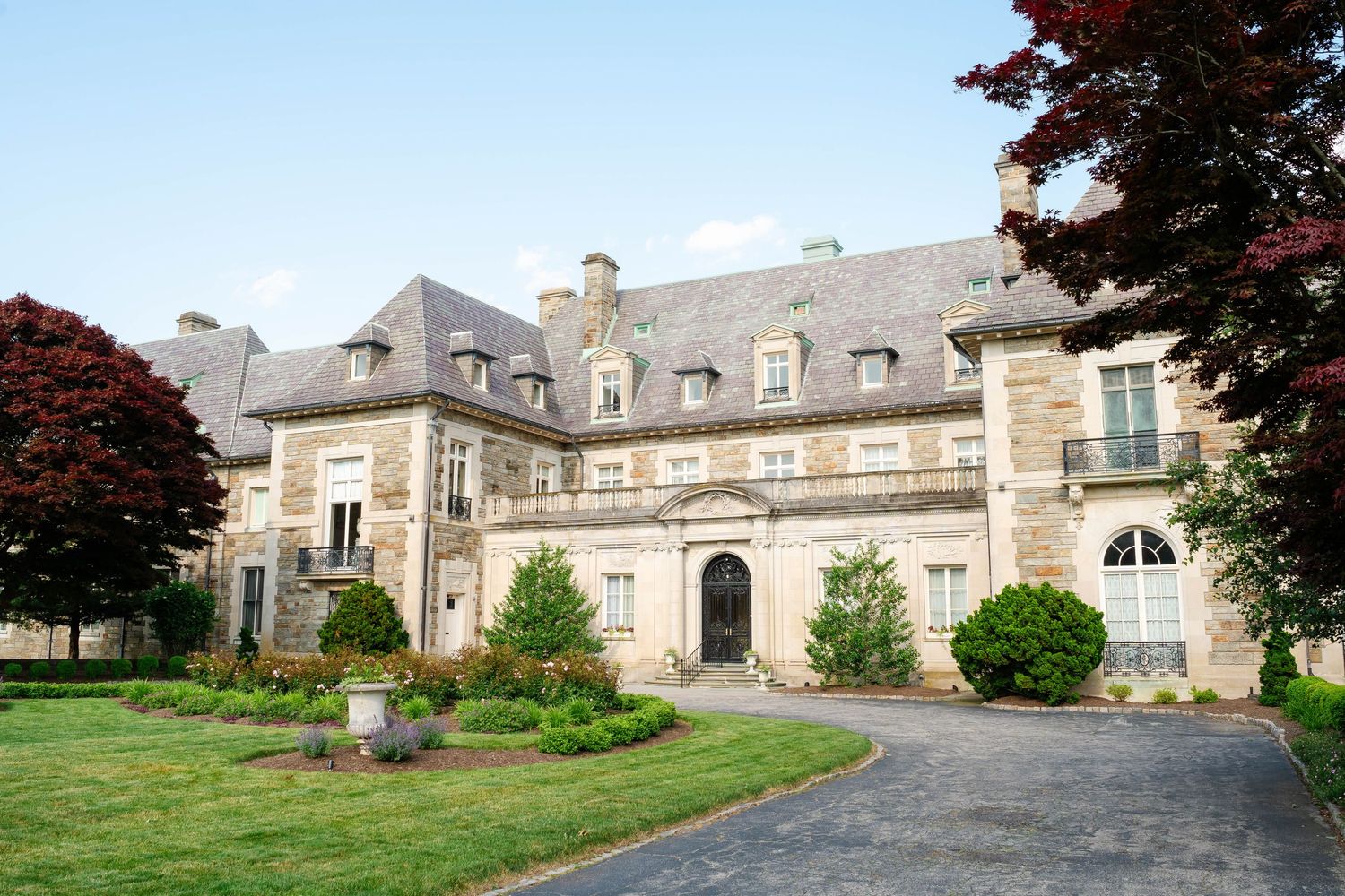 Grand stone manor house with manicured lawn and tree-lined driveway on a clear summer day.