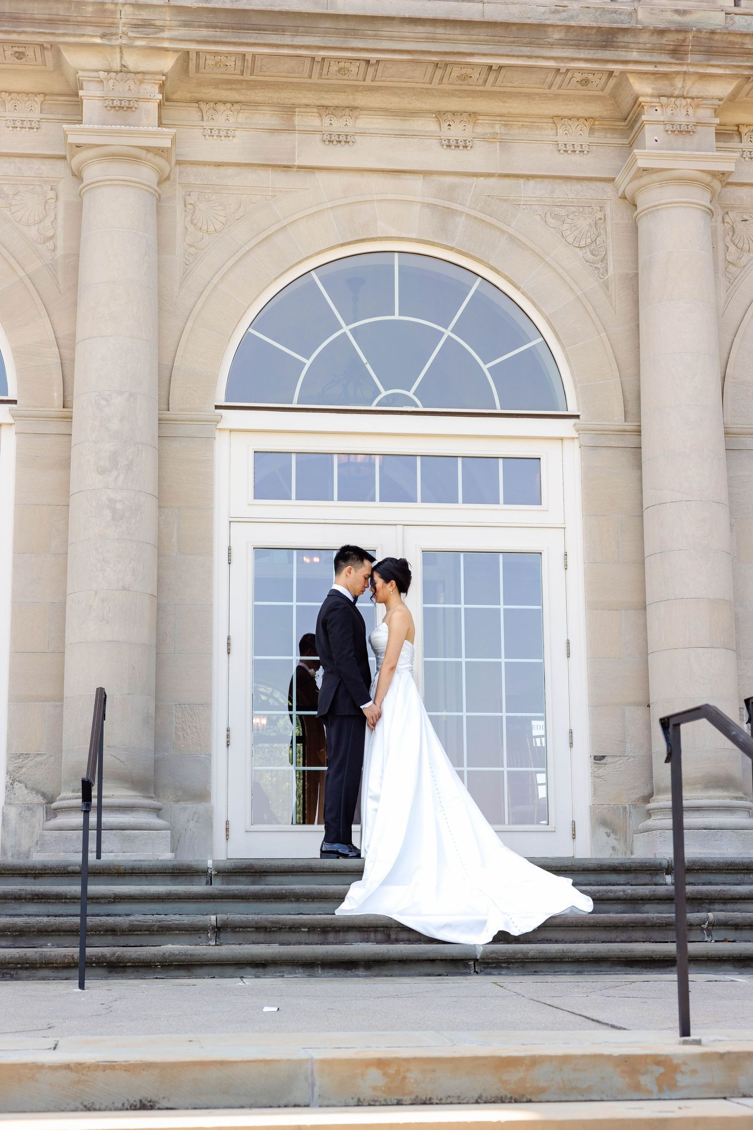 Bride and groom share a romantic moment on steps of elegant neoclassical building in wedding attire.