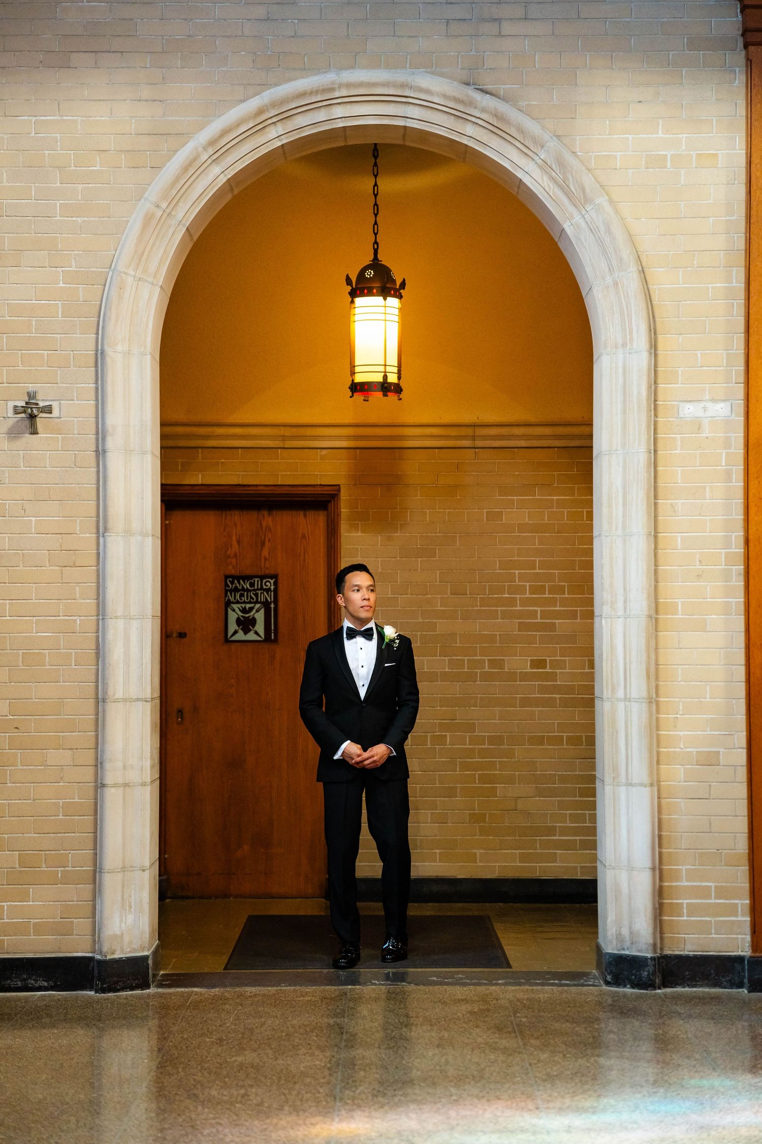 Groom in black tuxedo stands beneath stone archway with warm pendant light in church interior.