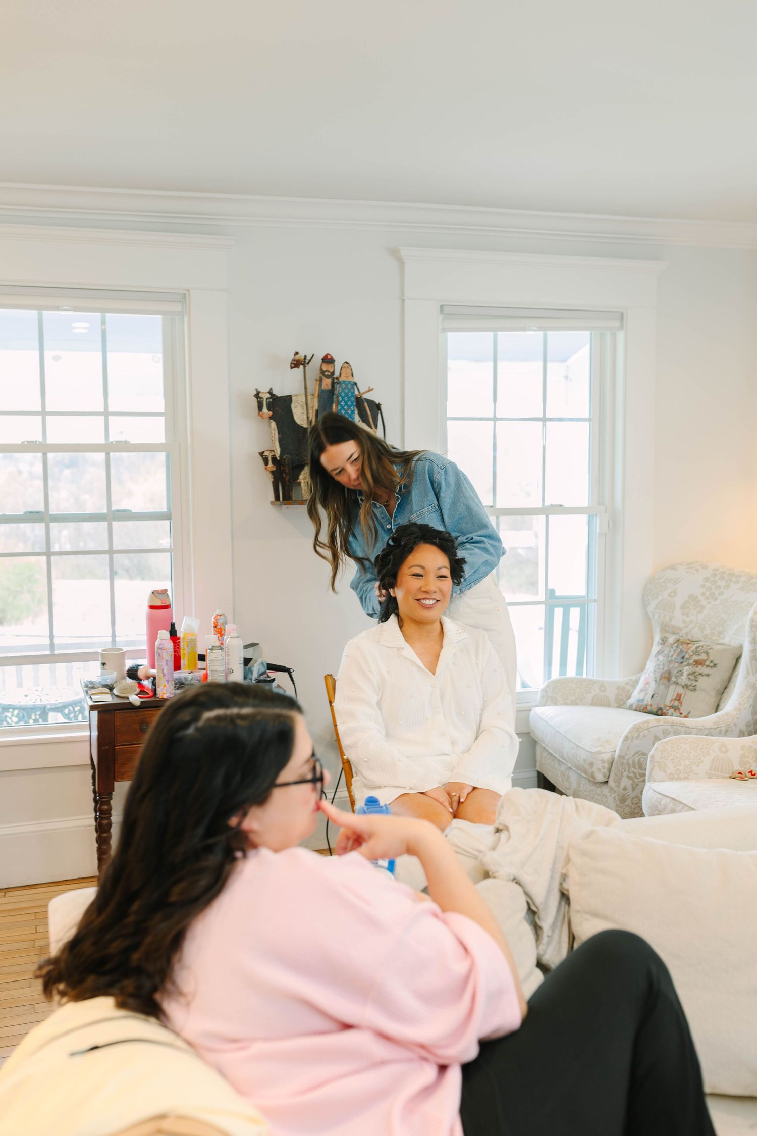 Bride smiles as hairstylist works on her hair in a bright white room while a bridesmaid relaxes nearby.