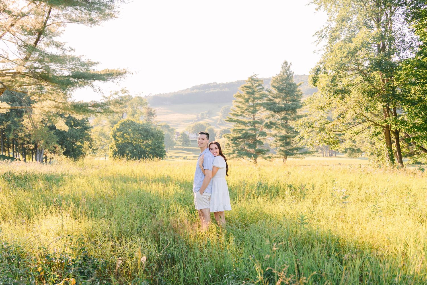 Two women in white dresses embrace in a sunlit meadow surrounded by lush green trees and rolling hills.