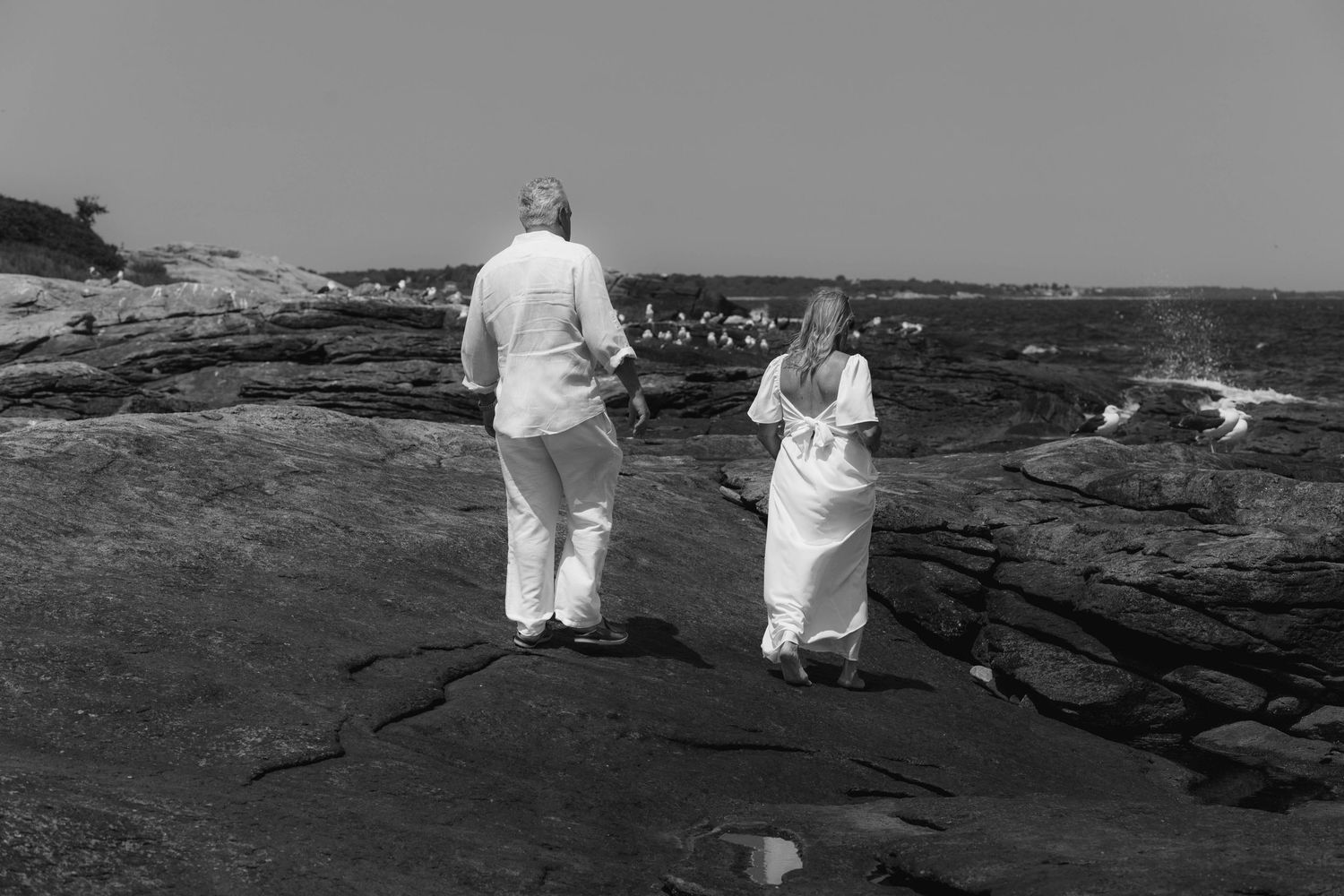 Two people dressed in white stand on rocky coastal terrain in a serene black and white photograph.