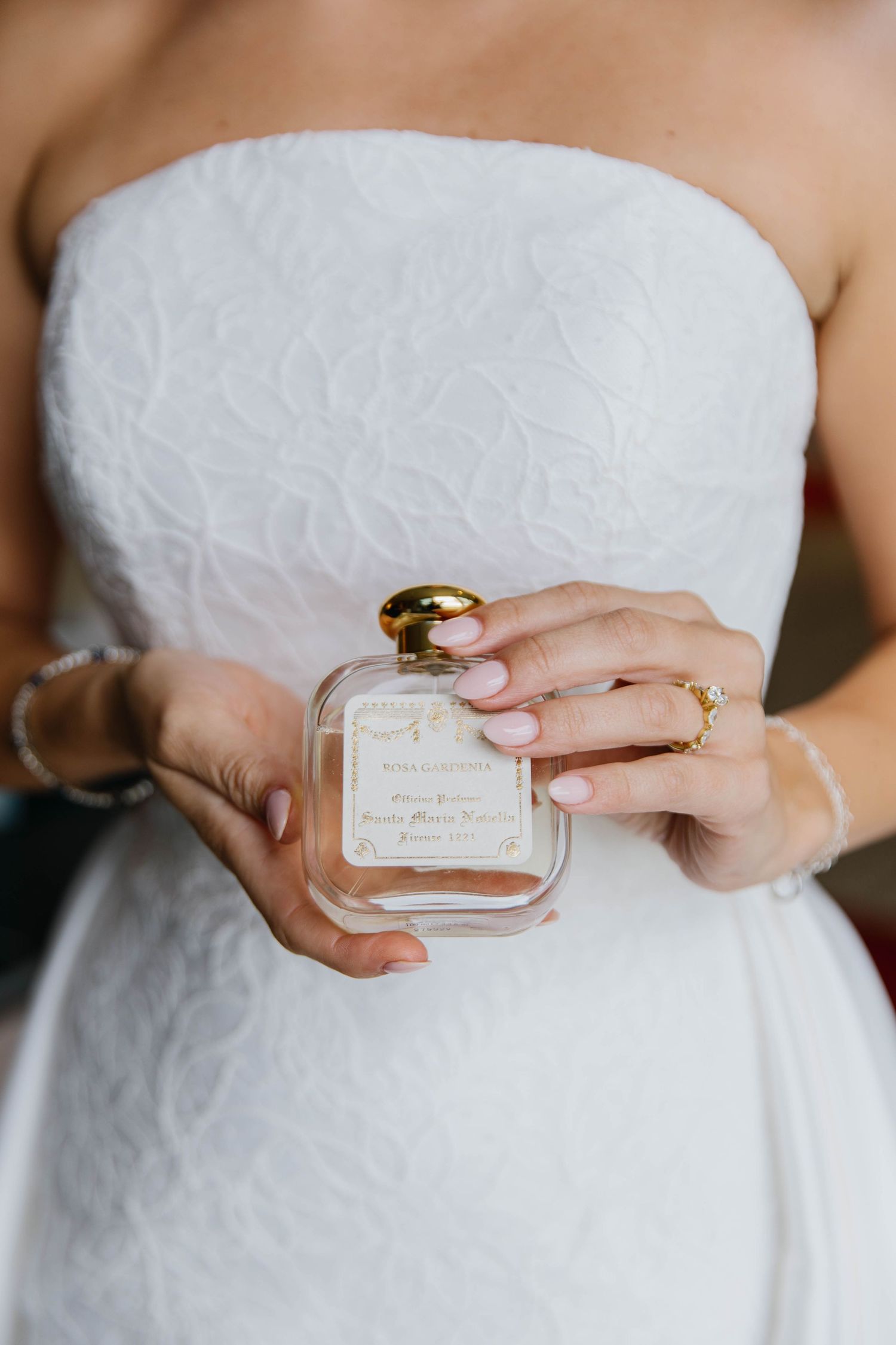 Bride in white strapless gown holding a personalized perfume bottle with gold cap and manicured nails.