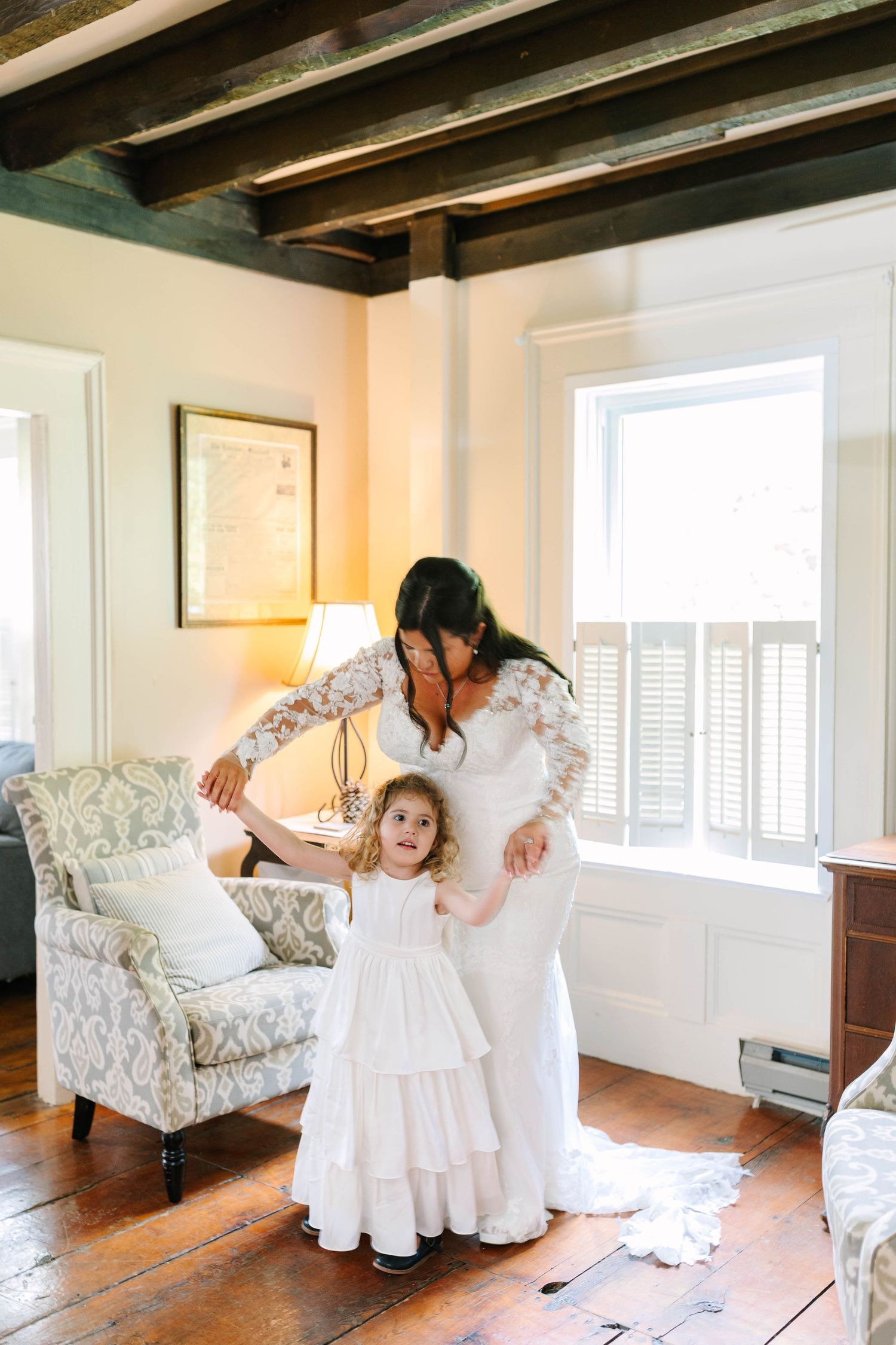 Bride in lace wedding dress helping flower girl near armchair indoors.