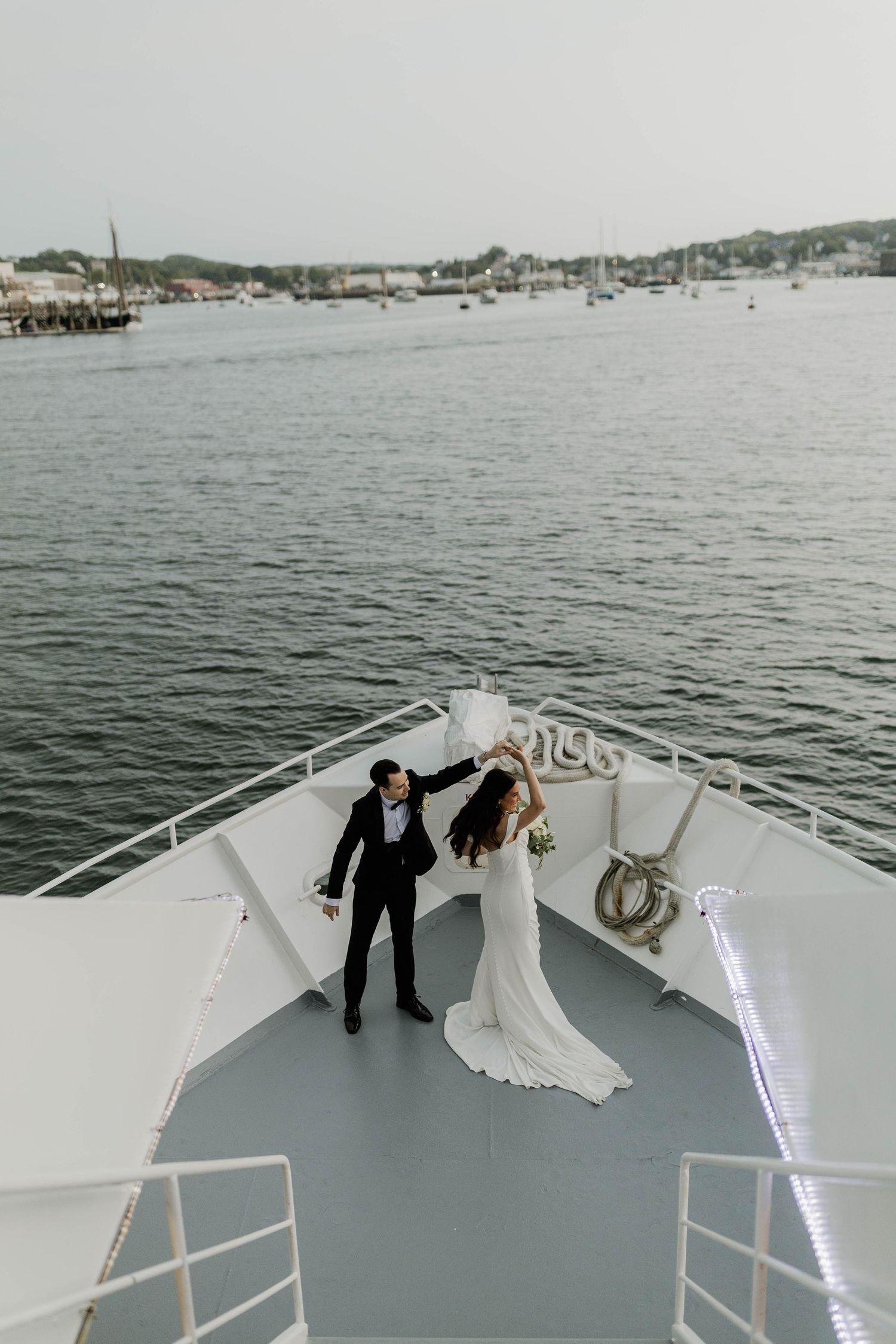 Groom twirling bride on the bow of a yacht, bride in white gown holding bouquet with harbor backdrop.
