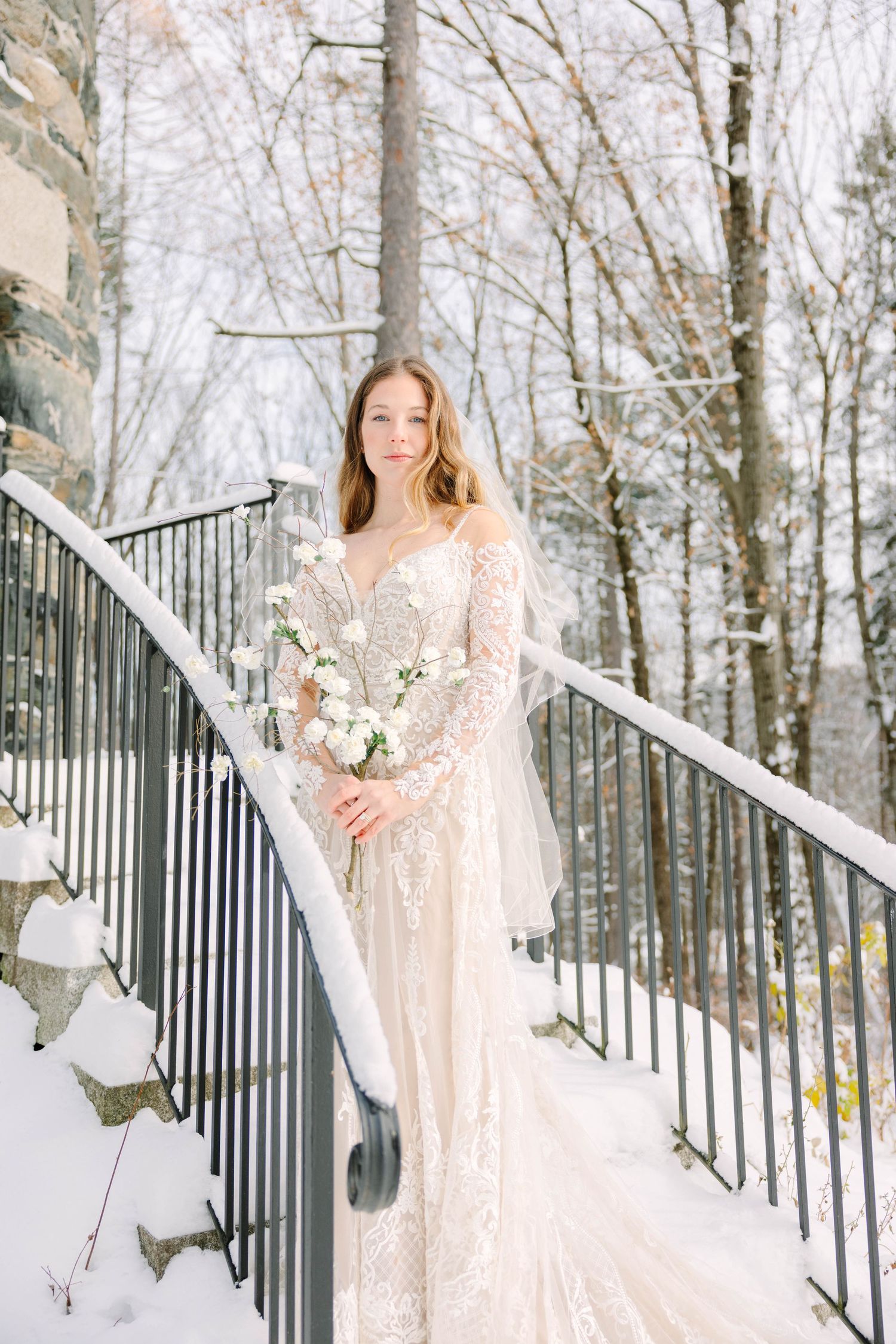Bride in long-sleeve lace gown holds white bouquet while standing on snow-covered outdoor staircase.