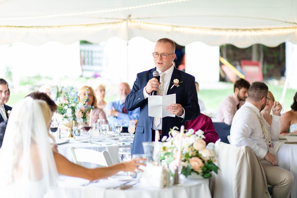 Father of the bride giving a speech with a microphone at an elegant tented wedding reception.