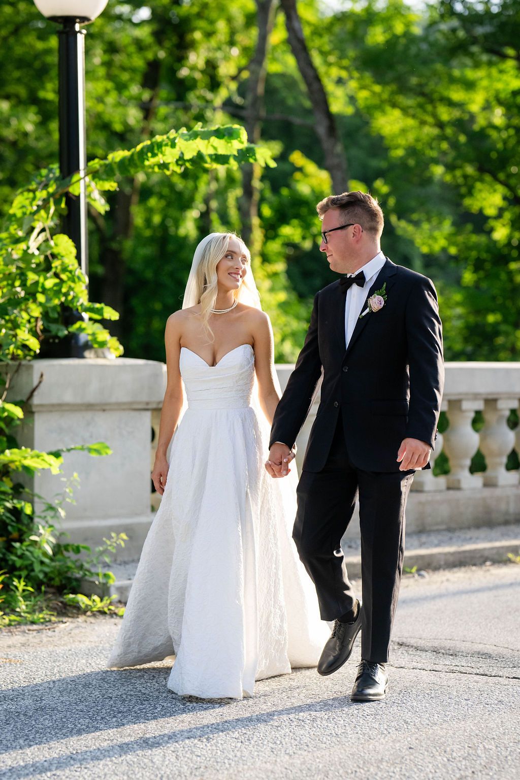 Smiling bride and groom in black tuxedo walk hand in hand on a sunlit garden path.