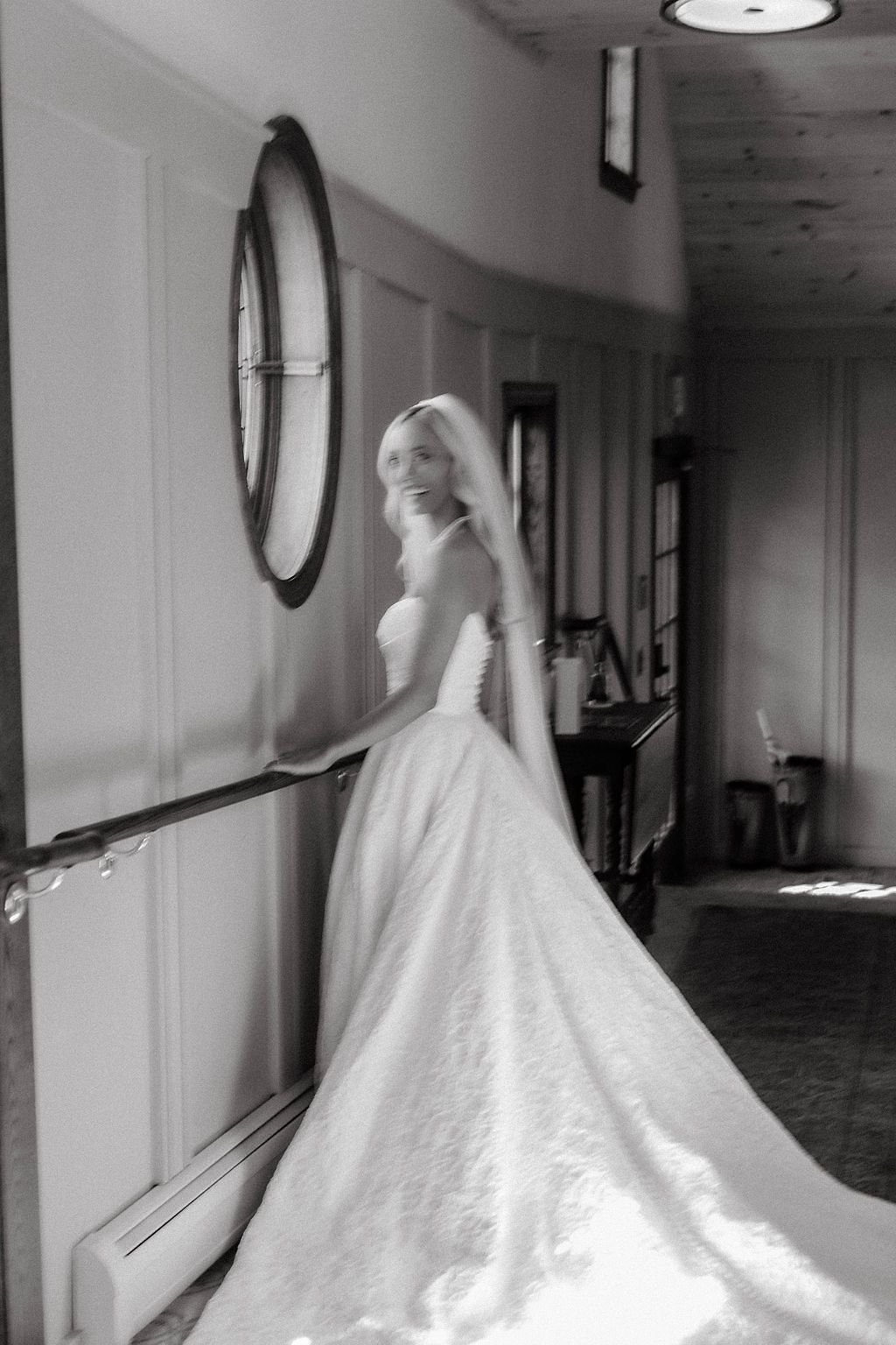 Black and white photo of a bride in a strapless gown and veil standing in a hallway.