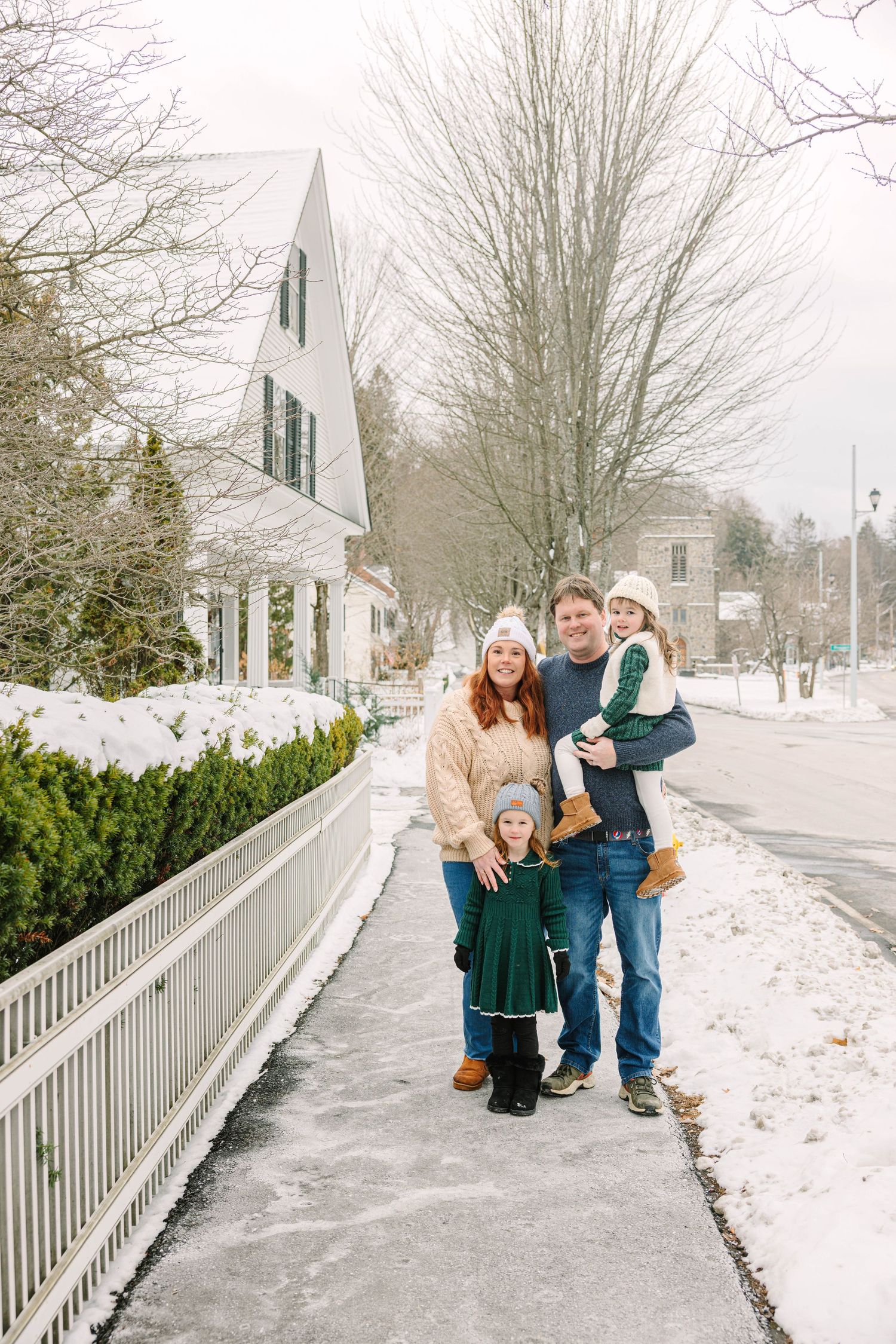 A smiling family of four poses together on a snowy sidewalk in a charming winter New England town setting.