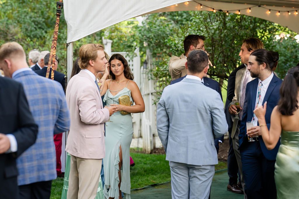 Wedding guests mingle and converse under a tent with string lights at an outdoor garden reception.