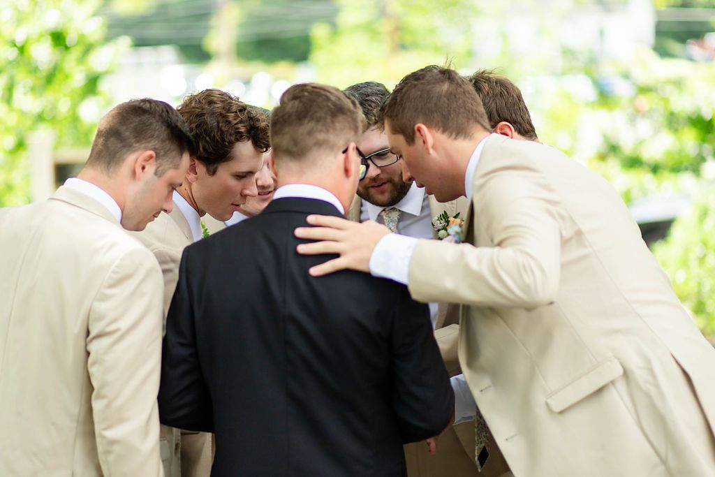 Groomsmen in tan suits gather in a circle with groom for a pre-wedding prayer or huddle outdoors.