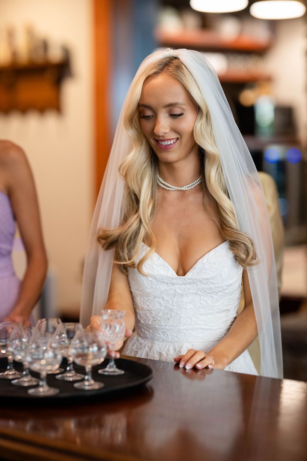 Smiling blonde bride in strapless lace gown and veil standing at bar with crystal glasses.