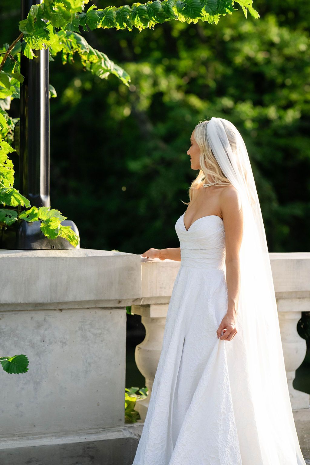 Bride in strapless white gown and cathedral veil stands beside a stone balustrade surrounded by lush green foliage.