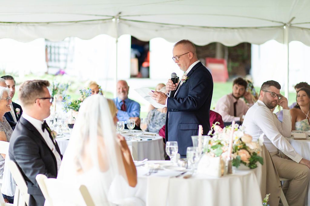 Father of the bride gives a touching toast speech at a sunlit tented wedding reception.