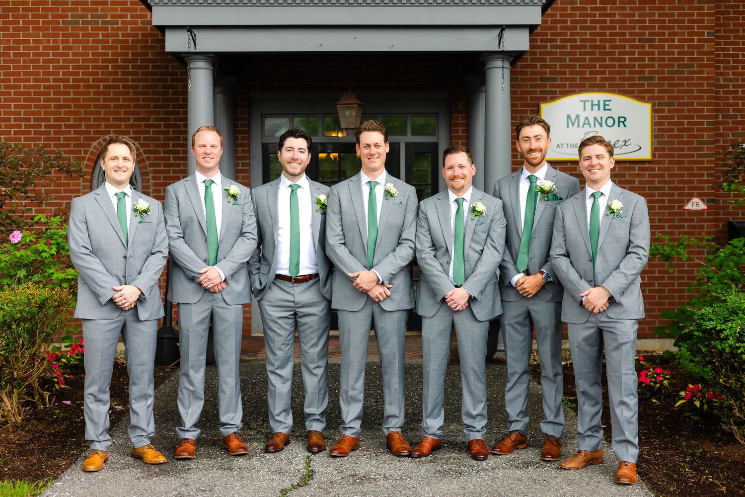 Seven groomsmen in matching gray suits and green ties pose outside The Manor at Essex.