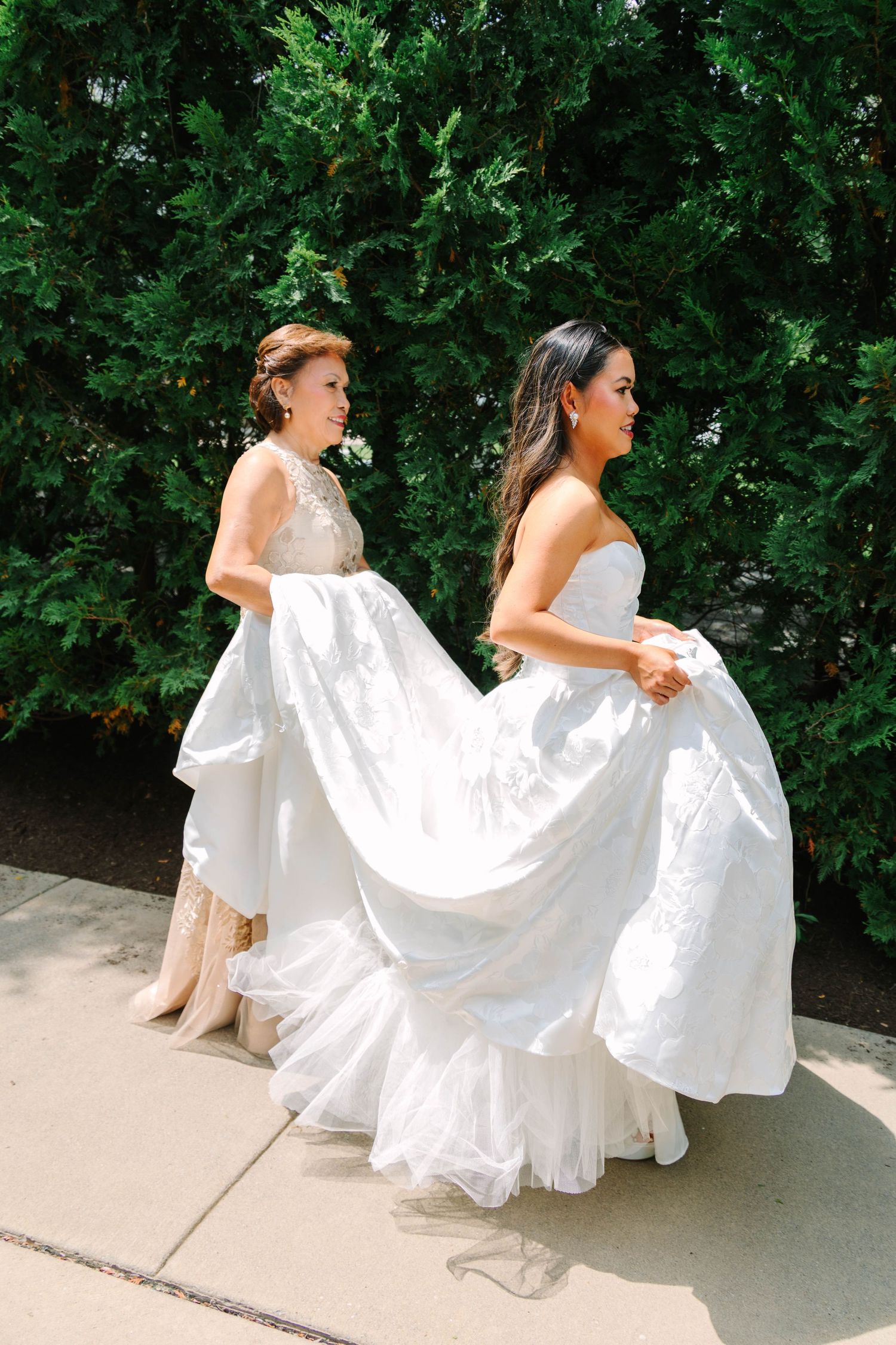 Bride in white strapless ballgown walking with helper lifting dress train outdoors.