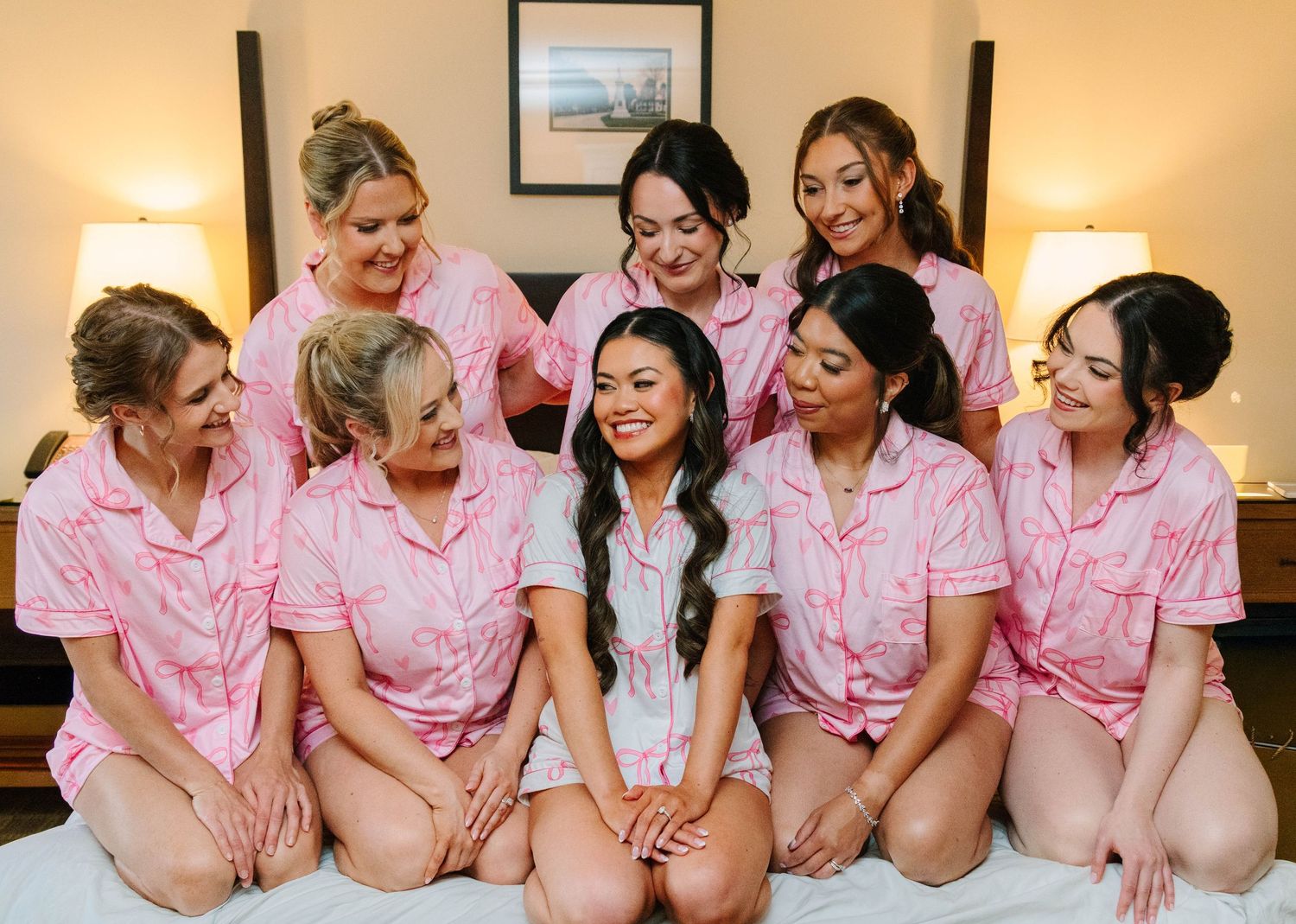 Bride and bridesmaids in matching pink pajamas smiling on hotel bed.