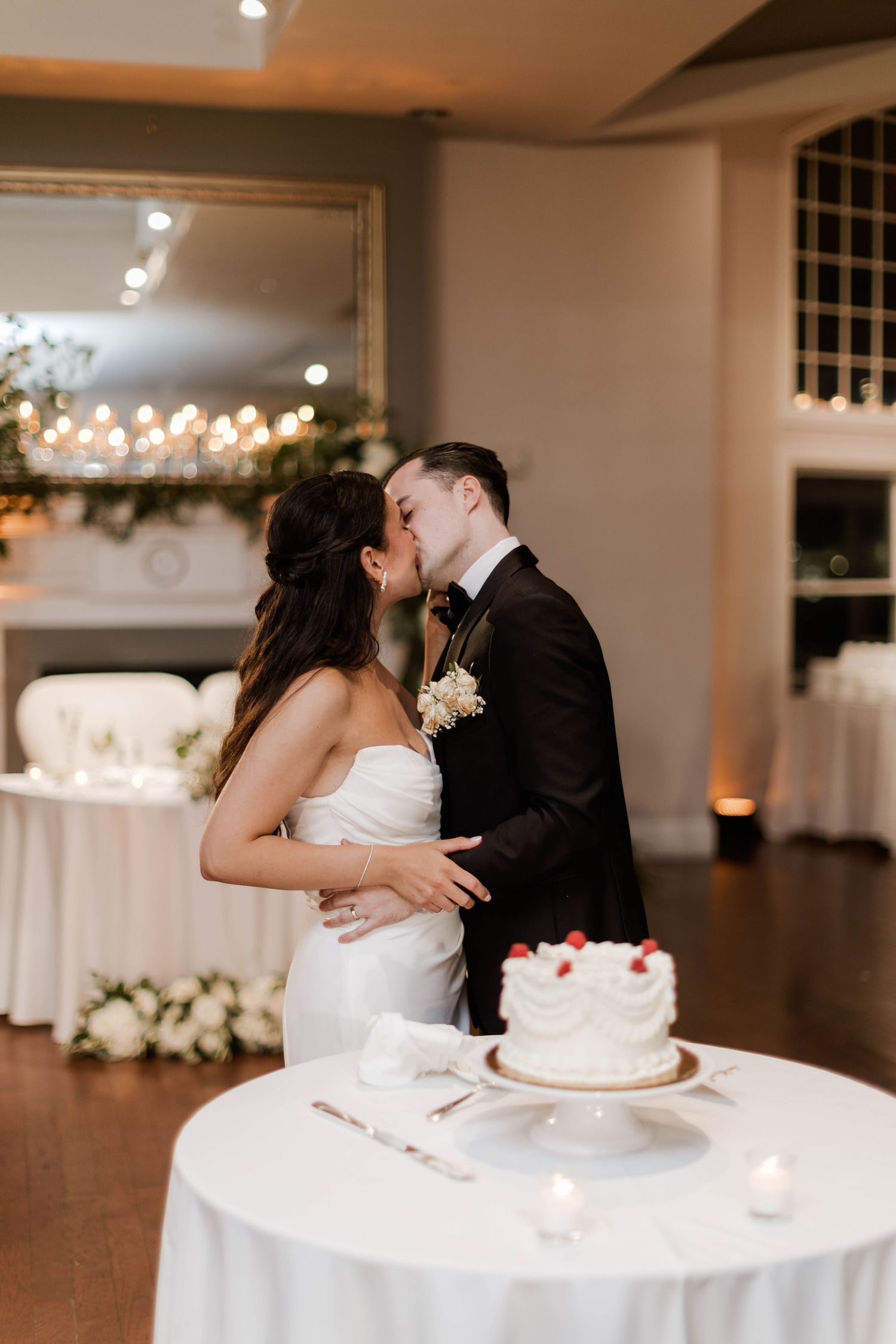 Newlyweds share a kiss beside their white strawberry-topped wedding cake at reception.