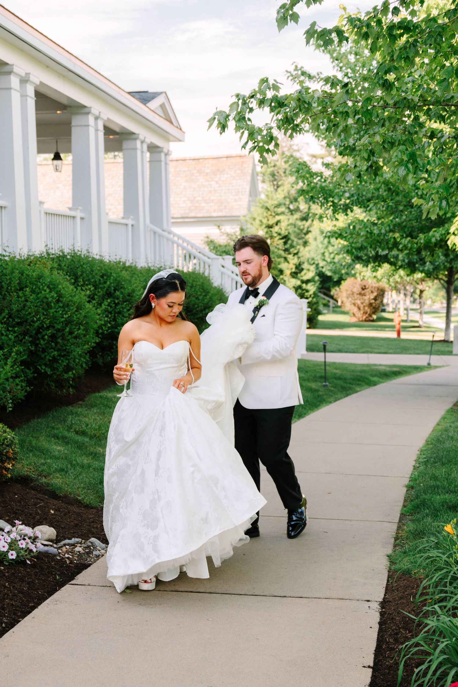 Bride in strapless lace ballgown walking with groom in white tuxedo jacket along garden pathway.