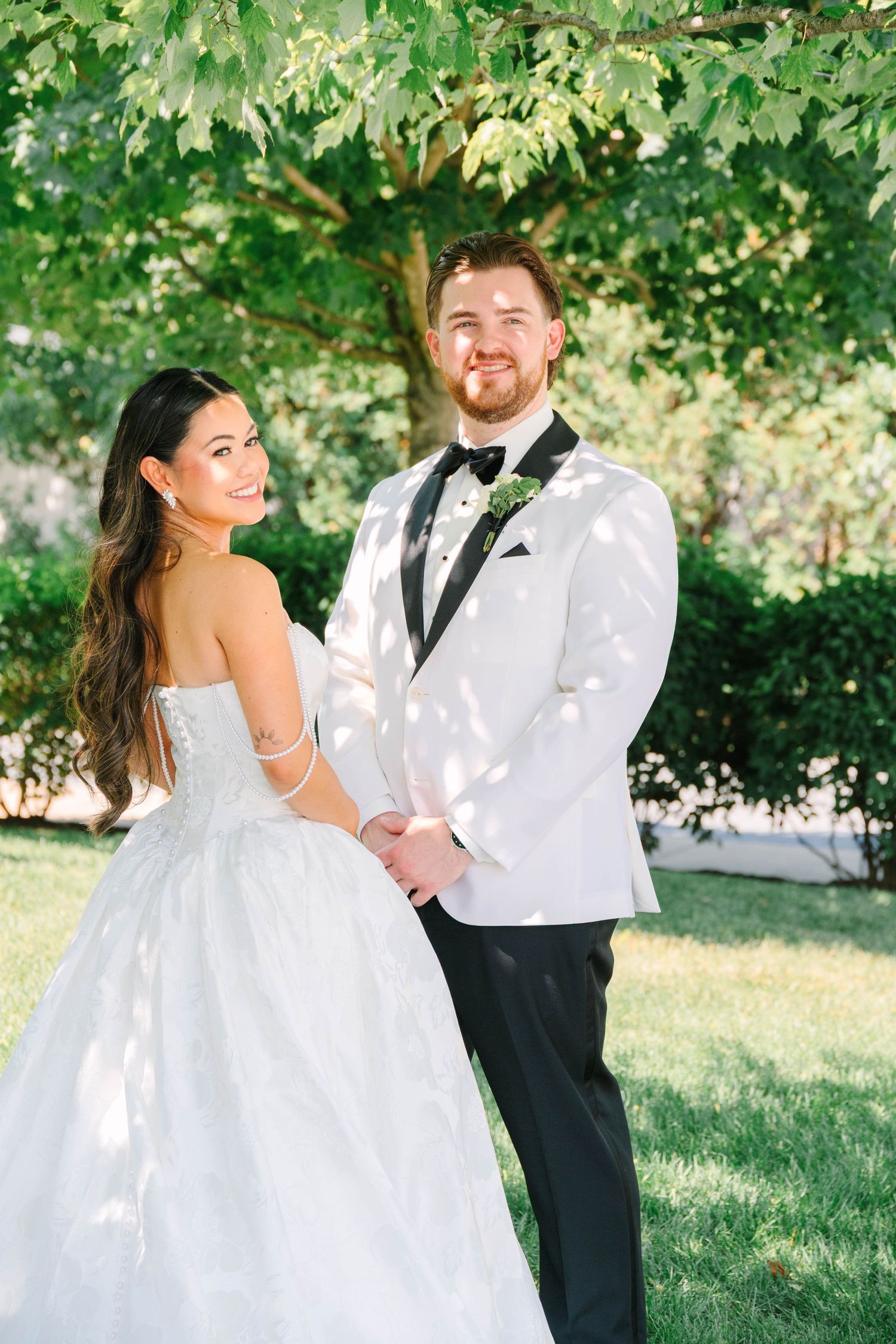 Bride and groom hold hands smiling under dappled sunlight in a lush garden setting.