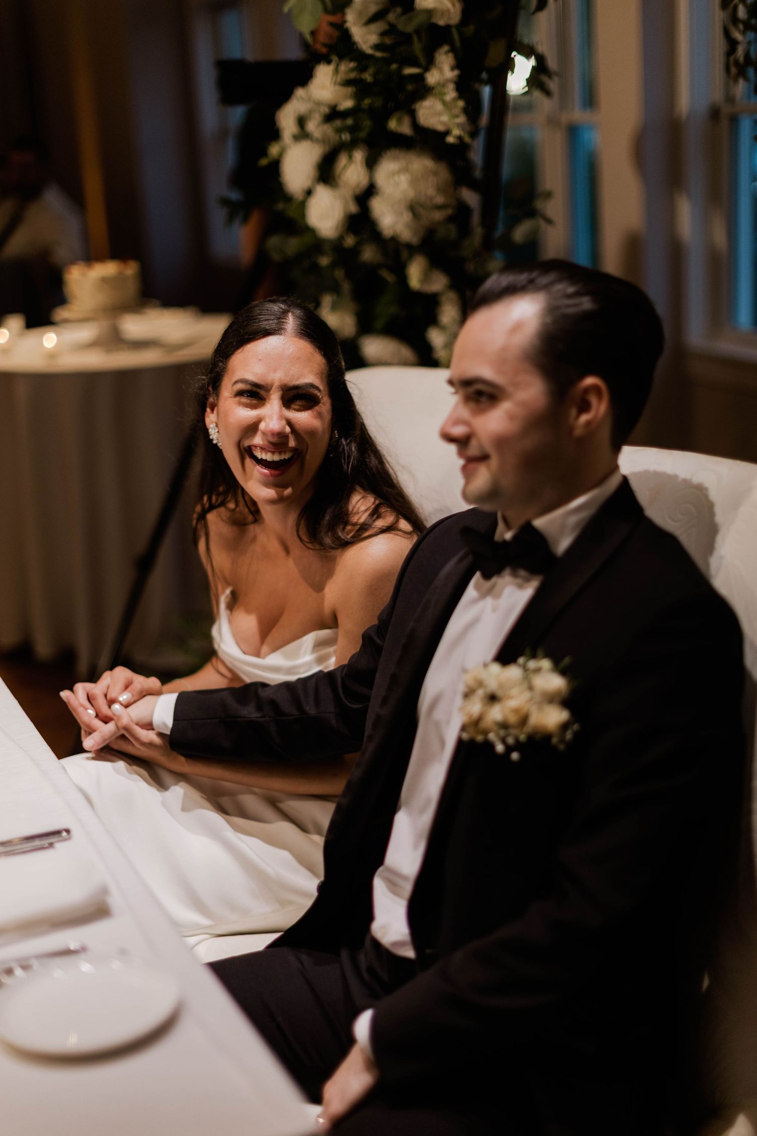 Bride laughing joyfully beside groom at sweetheart table during wedding reception dinner.