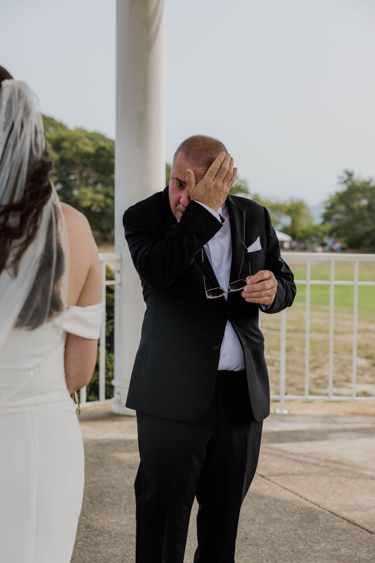 Emotional father wiping tears at wedding ceremony while holding glasses, dressed in black suit.