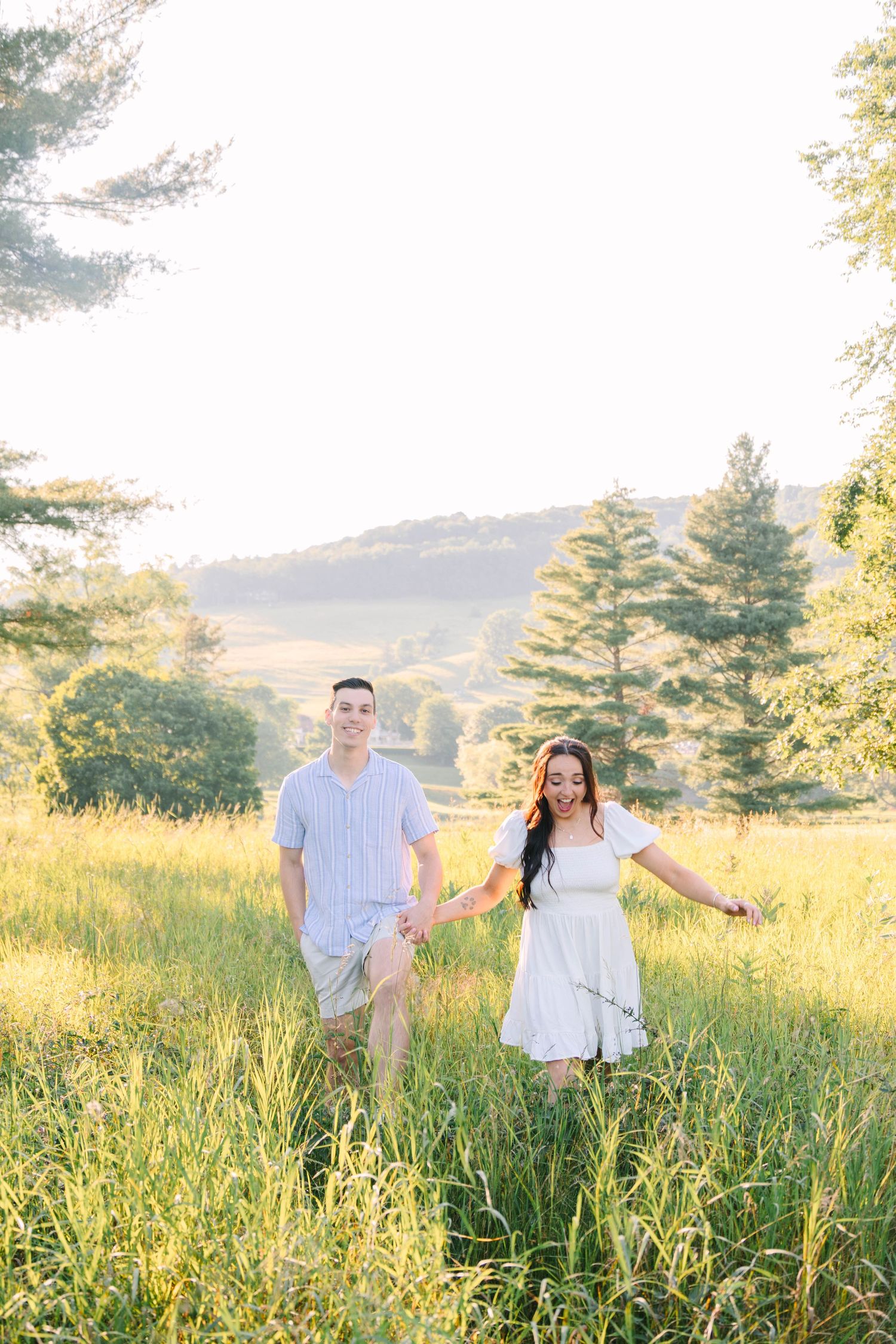 Couple holding hands walking through a golden sunlit meadow with rolling hills in background.