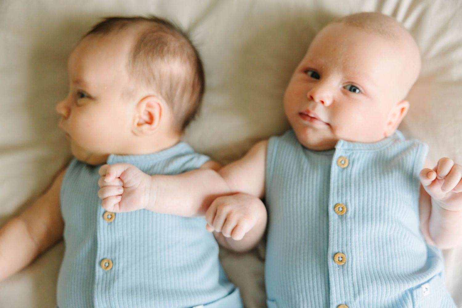 Two adorable twin babies in matching blue ribbed rompers lying together on a beige blanket.