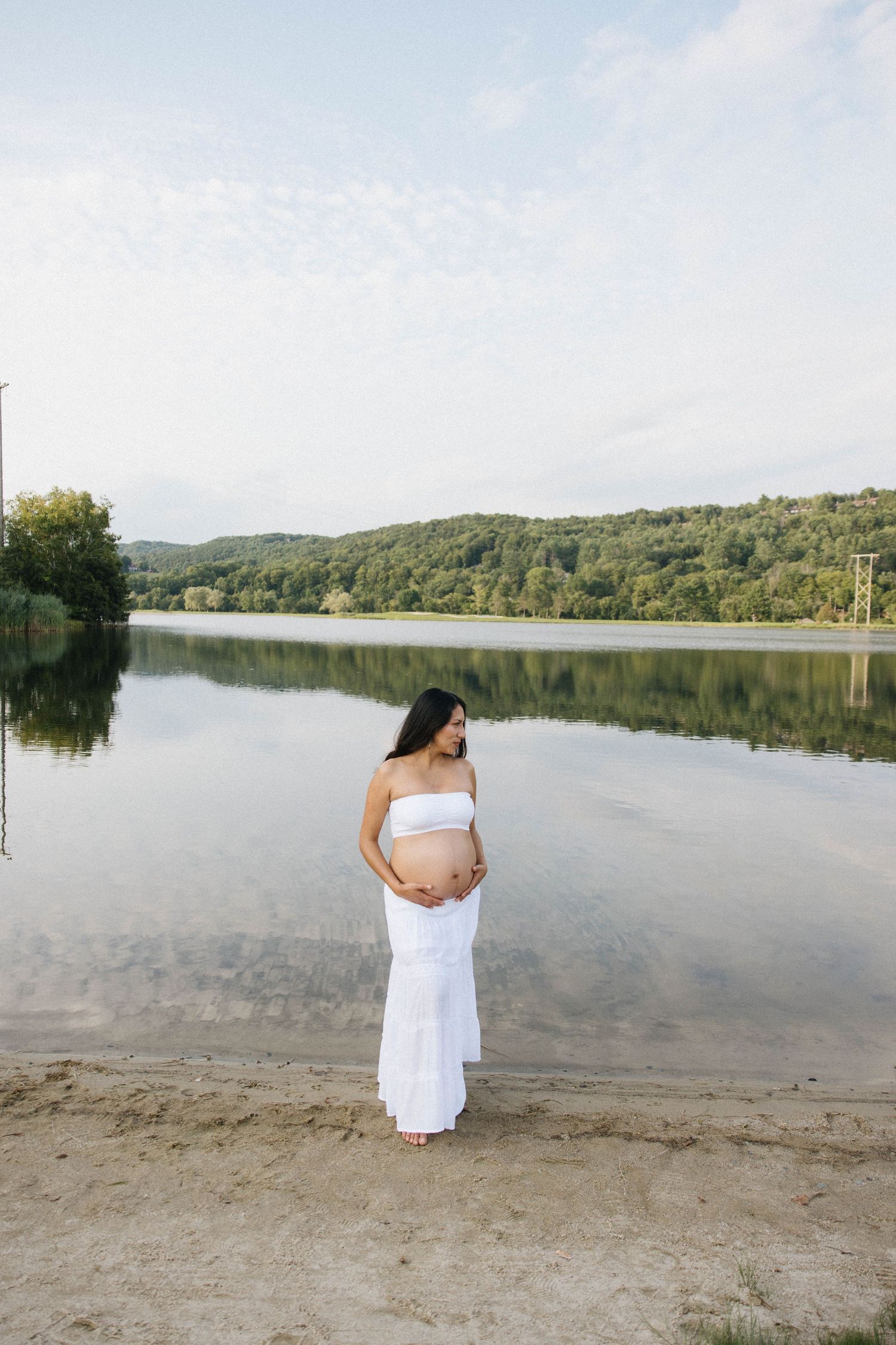 Pregnant woman in white outfit cradling baby bump by a calm lake with lush green hills reflected in water.
