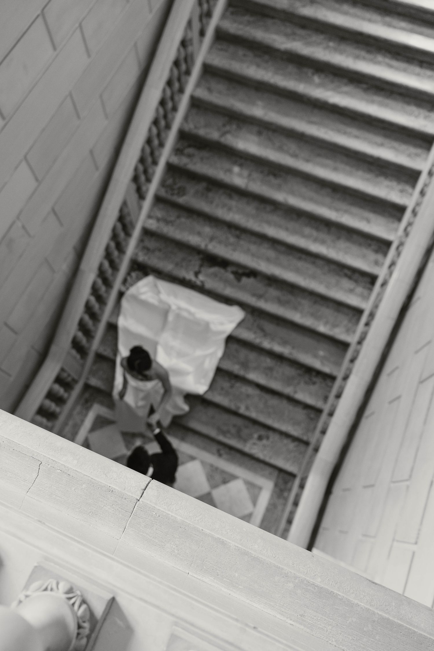 Black and white aerial view of bride and groom descending grand staircase with flowing wedding dress.