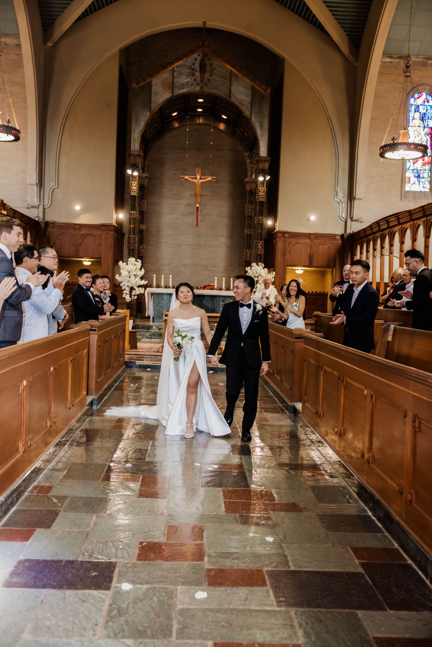 Bride and groom walking down church aisle after ceremony with guests applauding in Gothic-style chapel.