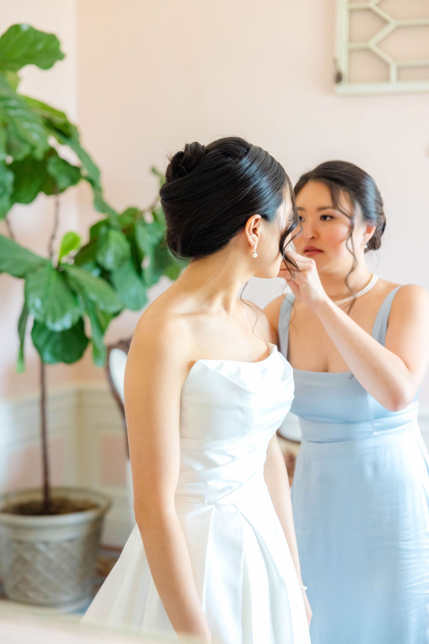 A bridesmaid in a light blue dress adjusts the hair of a bride in a white strapless gown before the wedding ceremony.