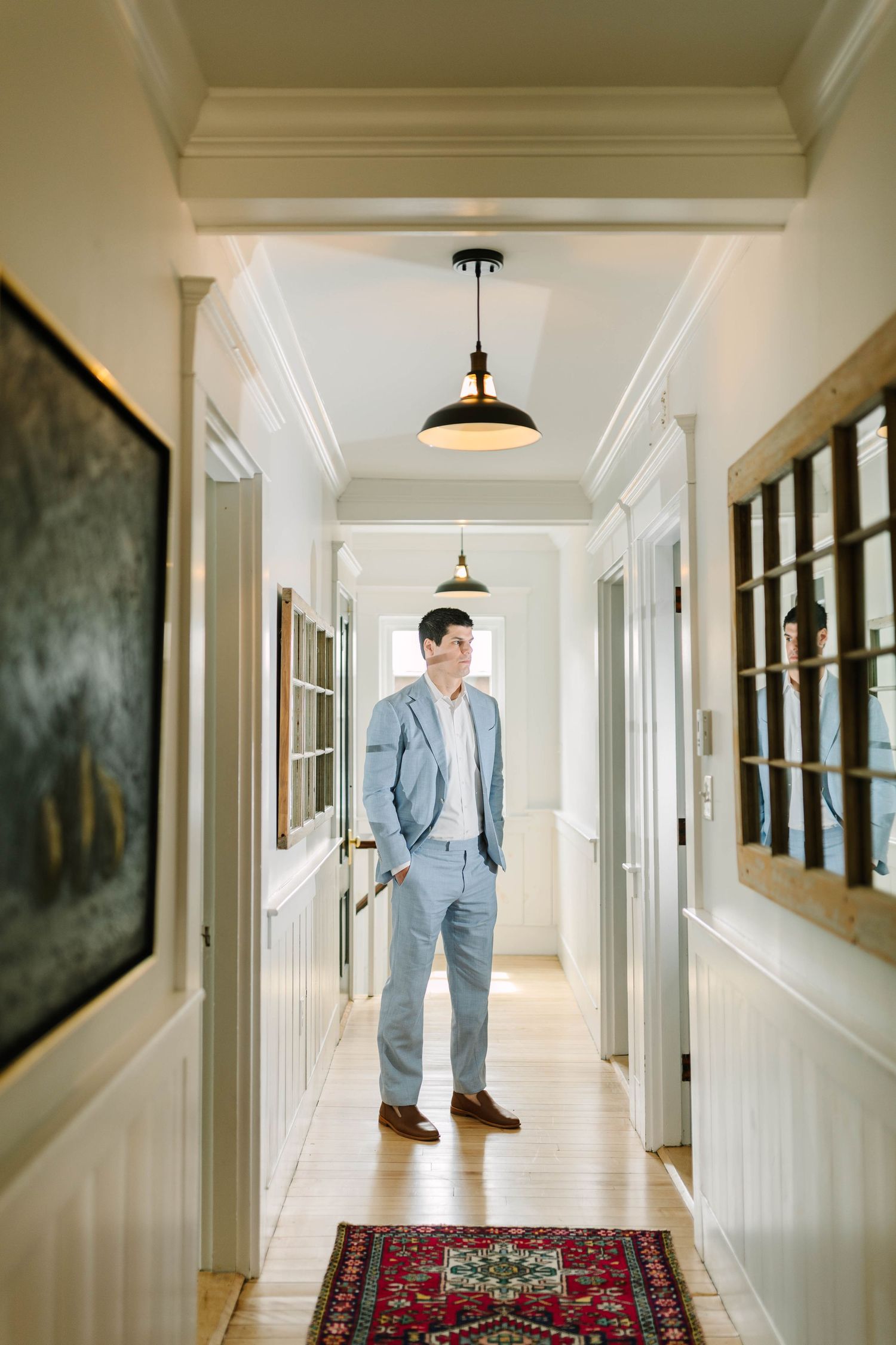 Groom in light blue suit standing in a bright white hallway with a red Persian rug.