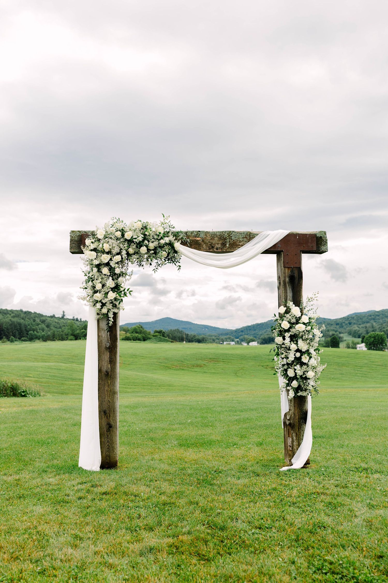 Rustic wooden wedding arch adorned with white roses and flowing fabric in a scenic green field.