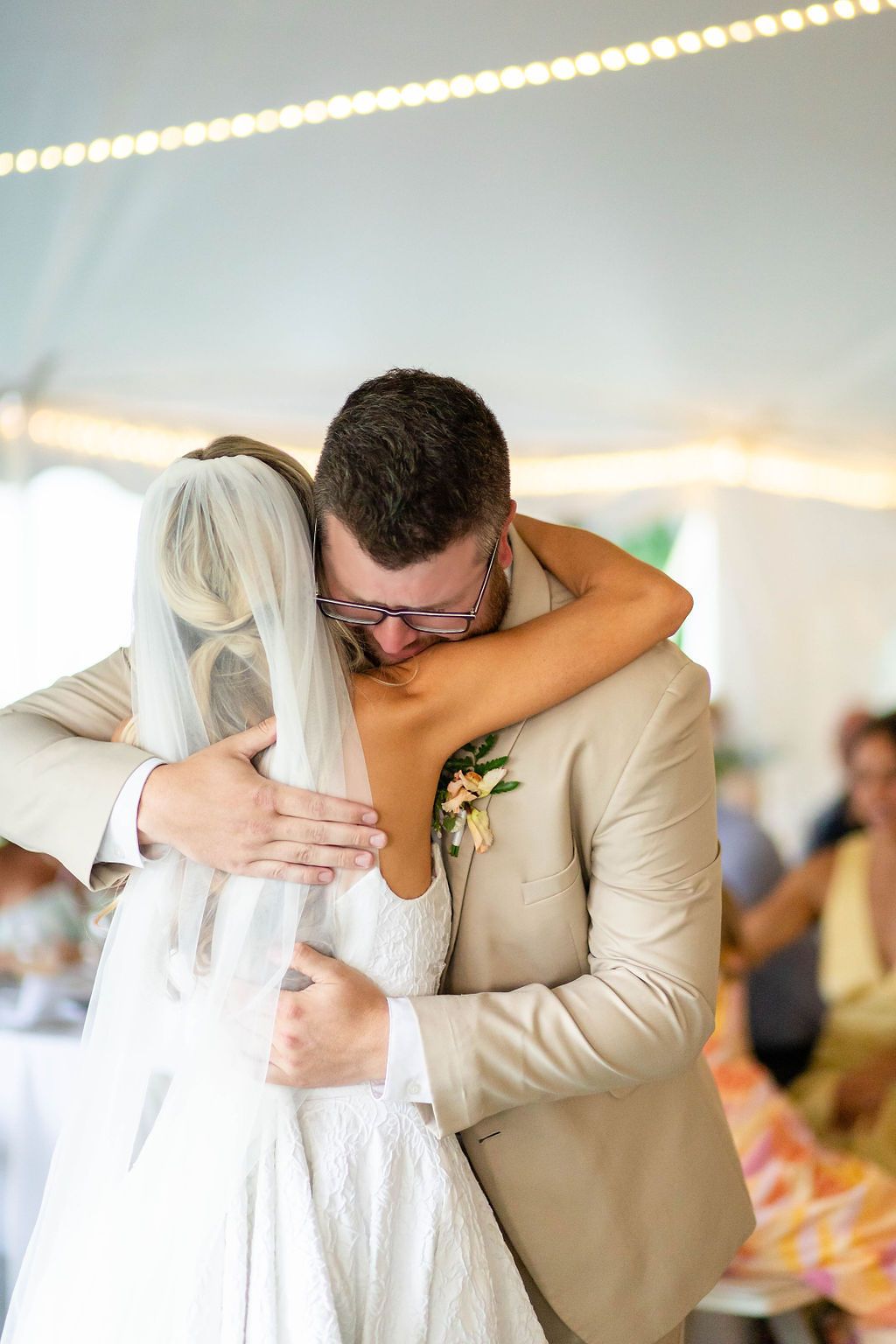 Emotional groom in tan suit embraces bride during their first dance under a white reception tent.