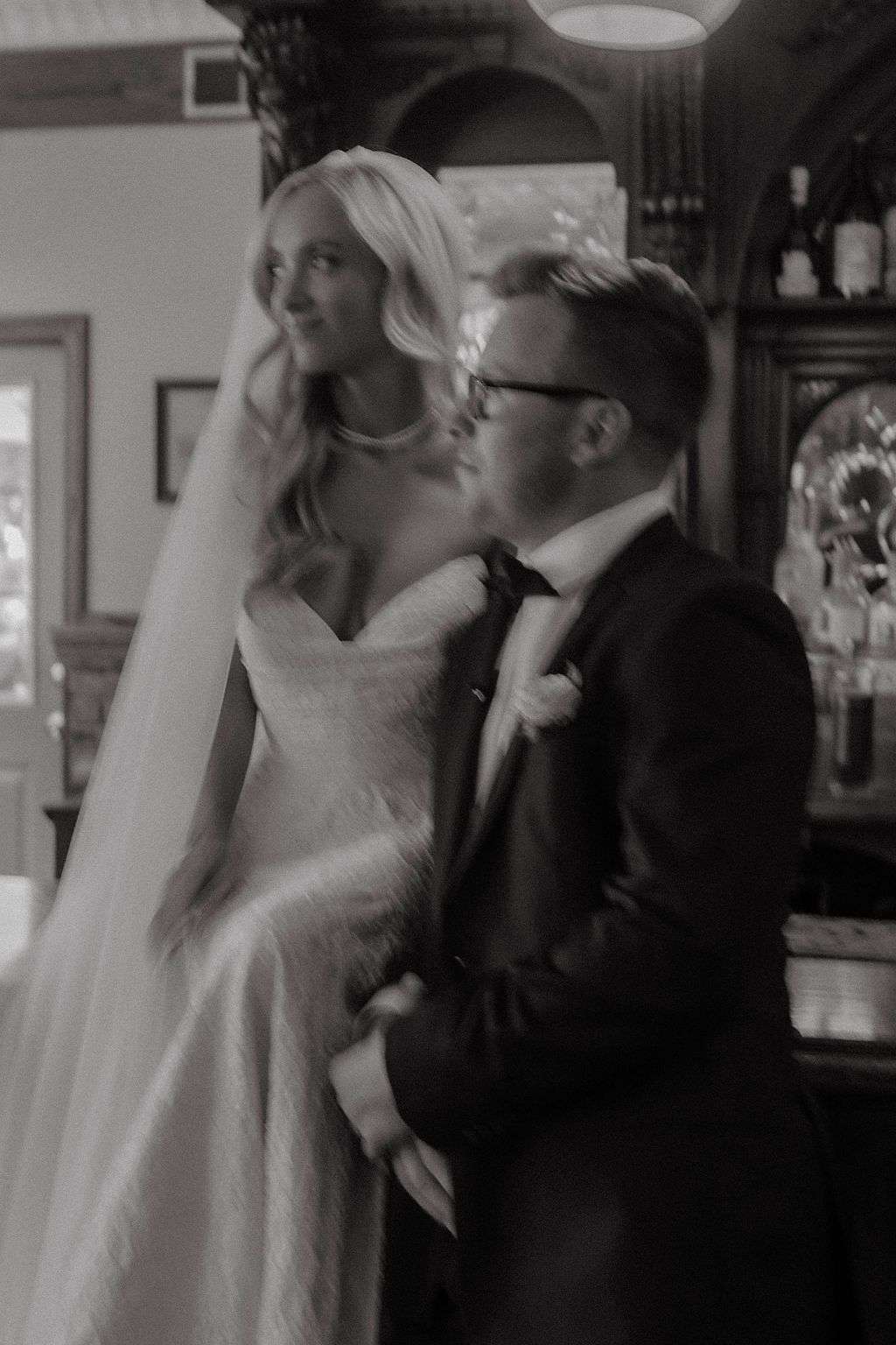 Black and white photo of bride in white gown and groom in tuxedo sharing an intimate moment at a bar.