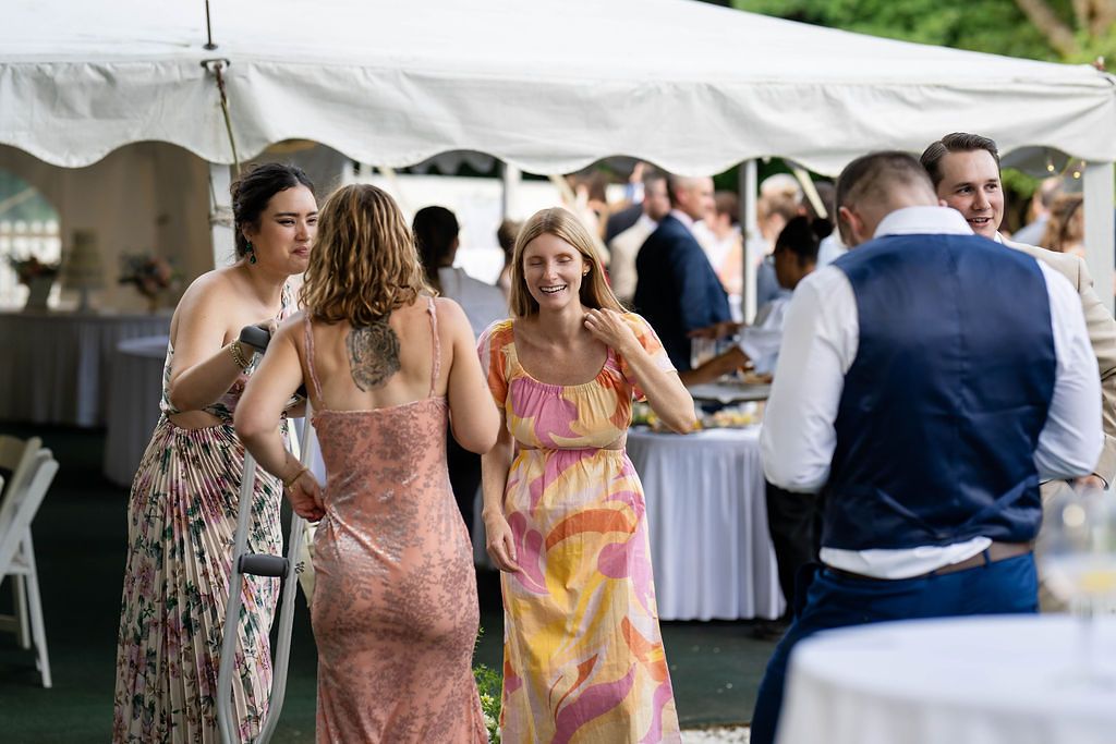 Wedding guests in colorful summer dresses mingle under a white tent at an elegant outdoor wedding reception.