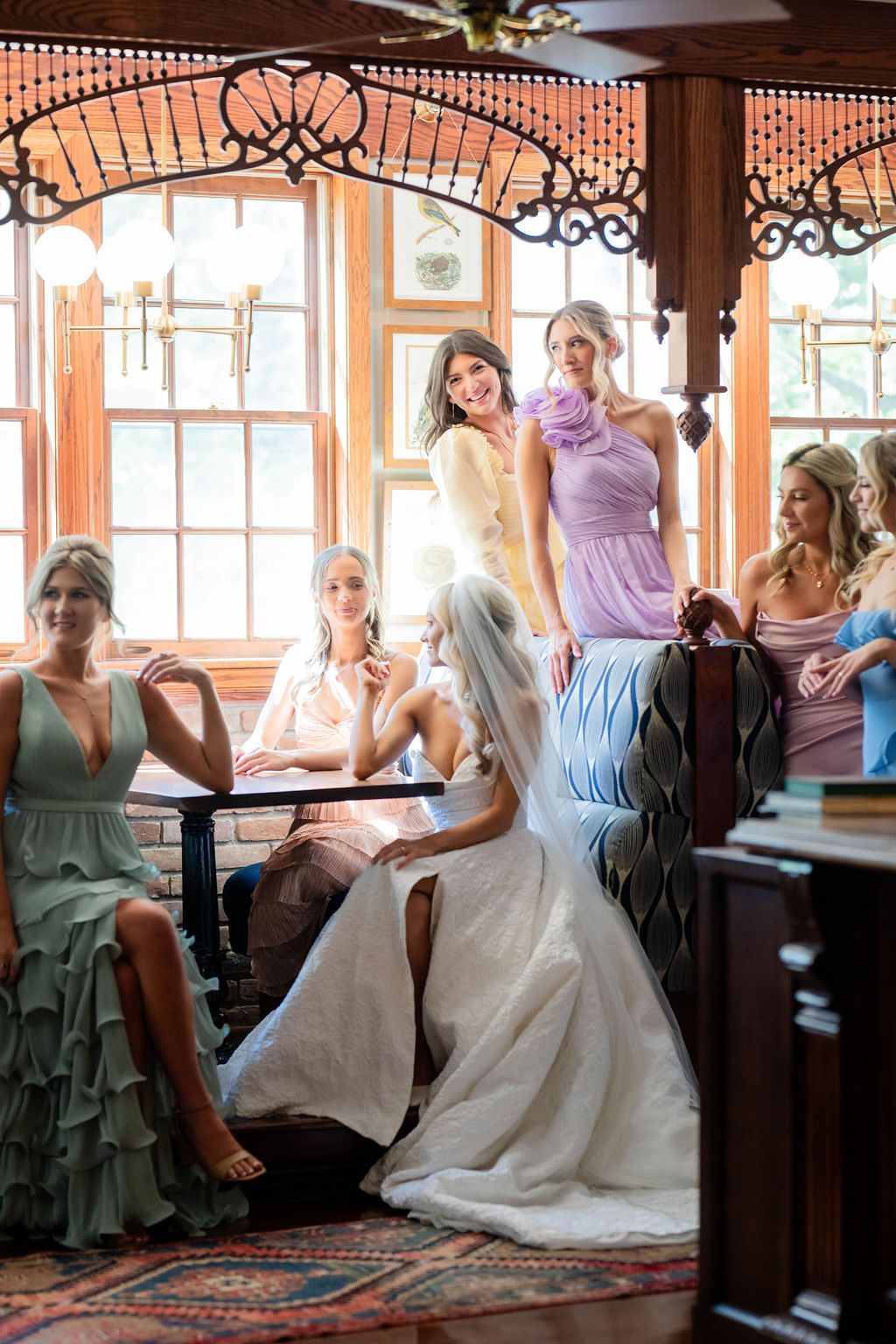 Bride surrounded by bridesmaids in pastel gowns inside an elegant vintage venue with ornate woodwork.