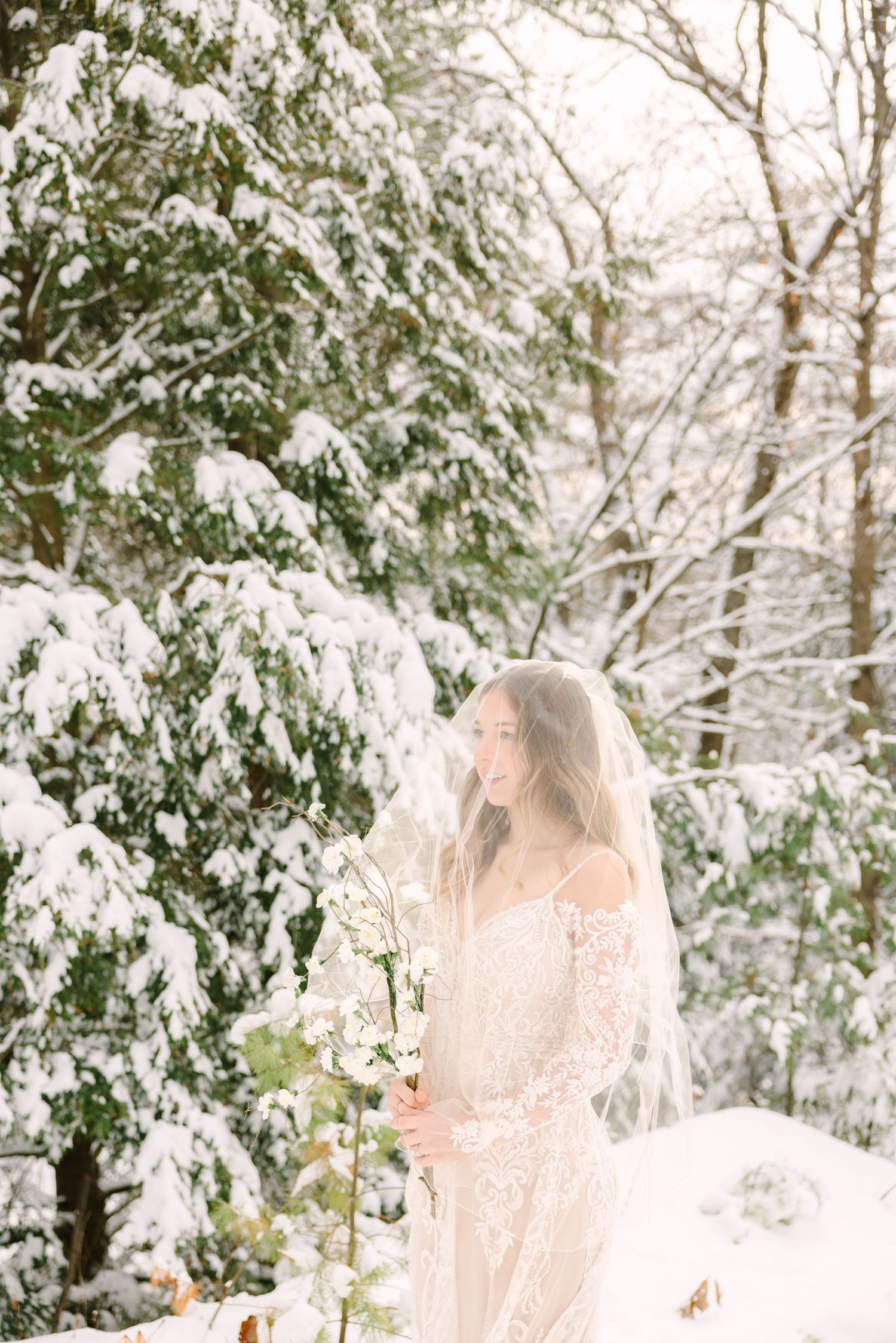 Bride in lace wedding dress and veil smiles softly holding white flowers near snow-laden evergreen trees.