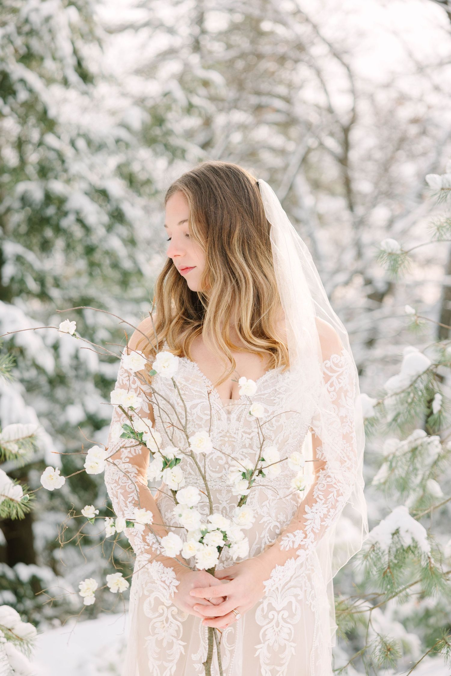Bride in lace gown and veil holds white floral bouquet among snow-covered evergreen trees in winter.