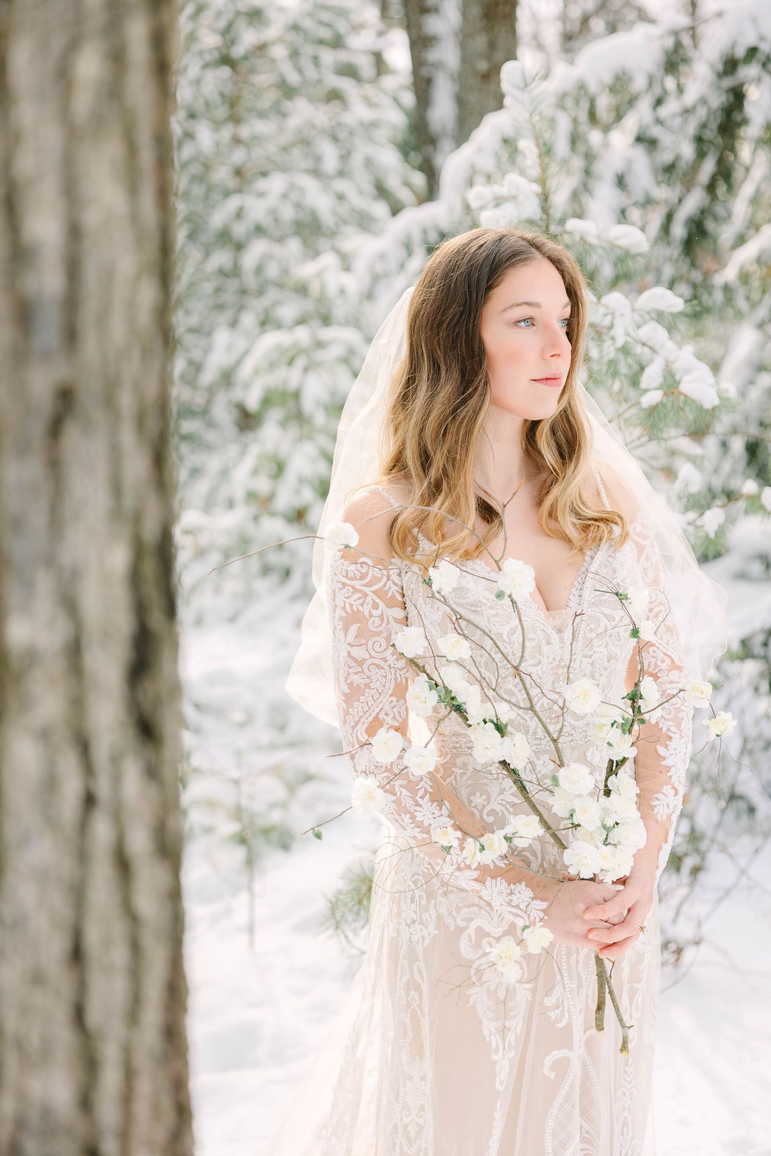 Elegant bride in lace dress with veil gazes aside holding white blooms in snowy winter woodland setting.