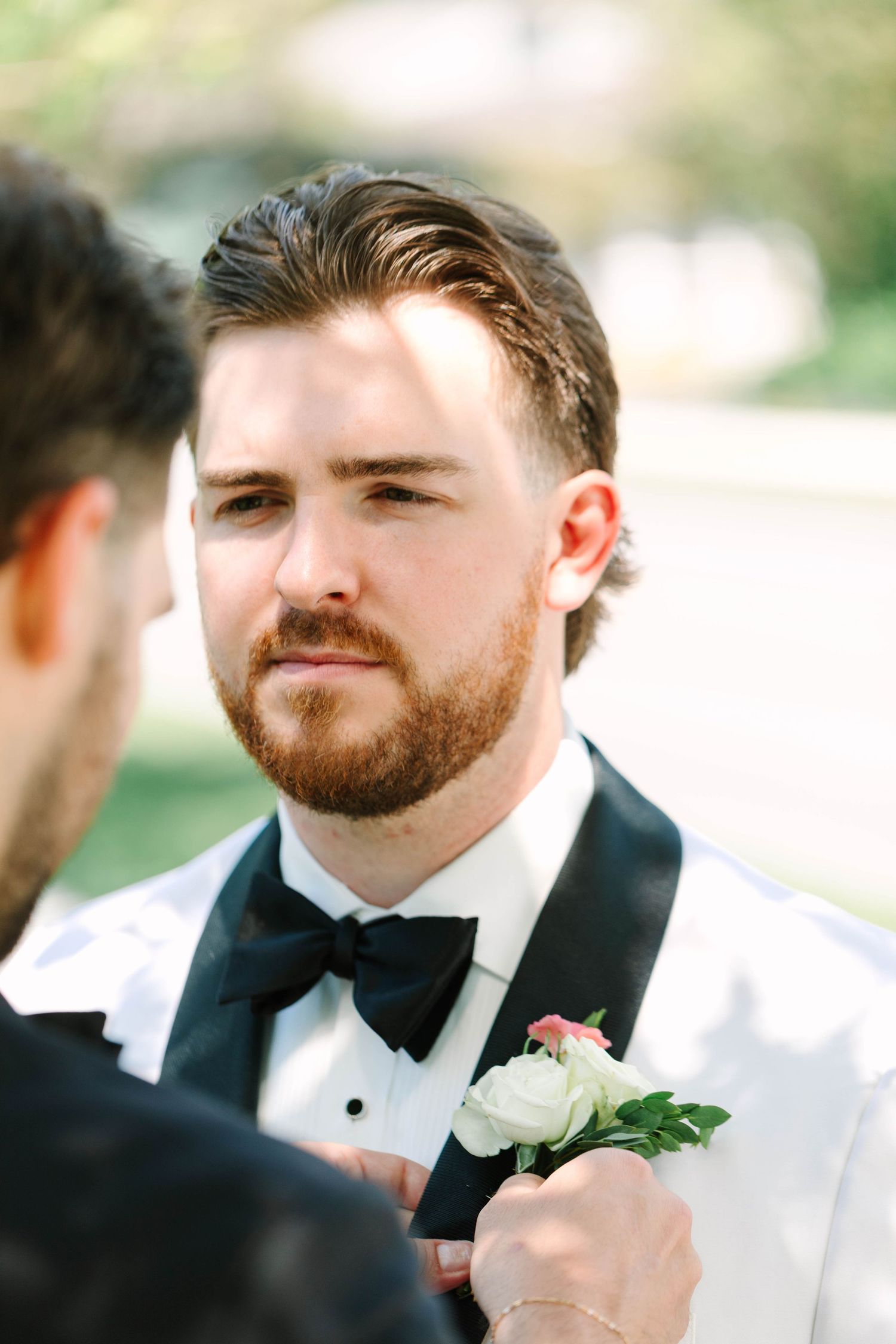 Groom in white tuxedo having boutonniere pinned to lapel outdoors before wedding ceremony.