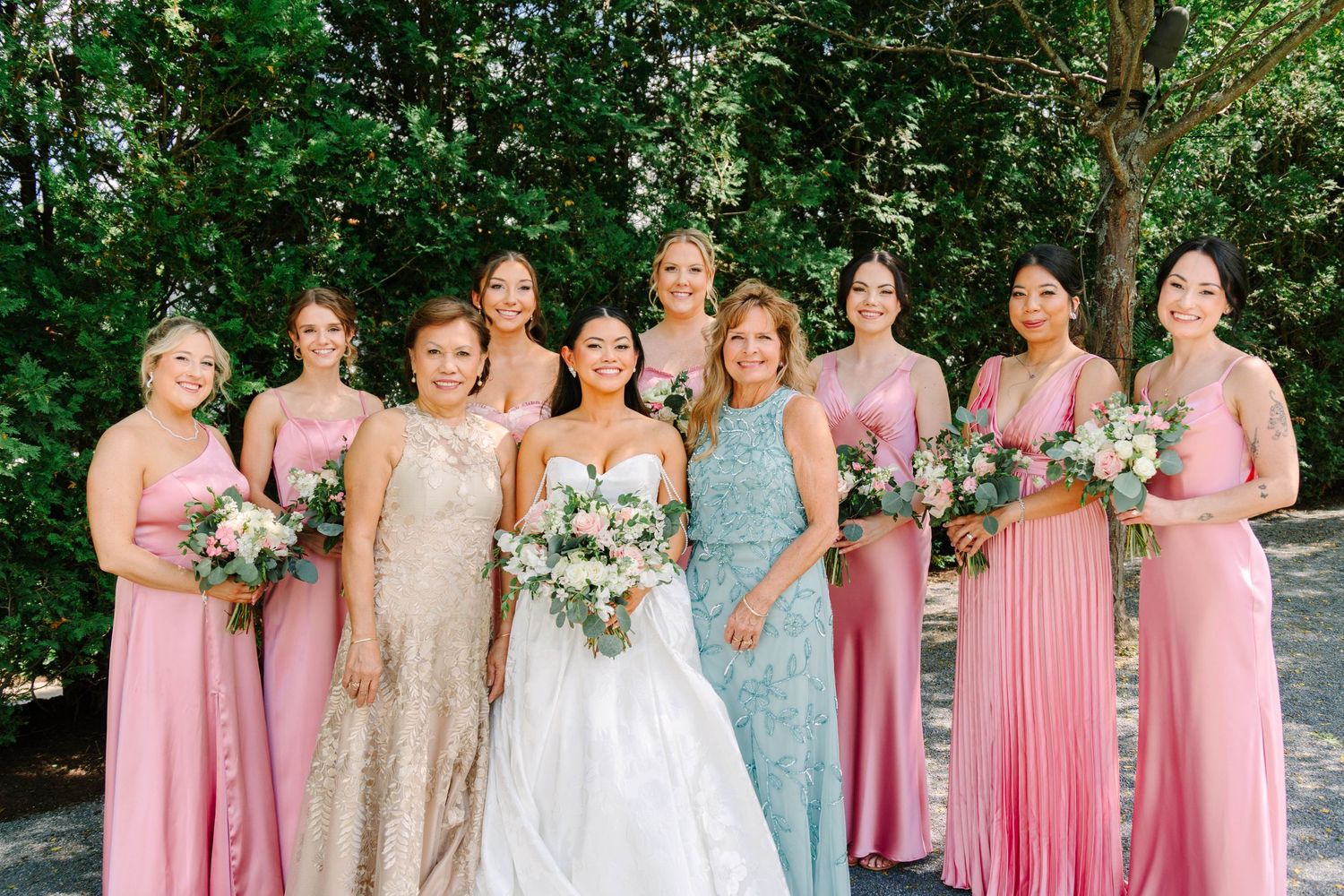 Bride in white gown poses with bridesmaids in mixed pink dresses and floral bouquets outdoors.