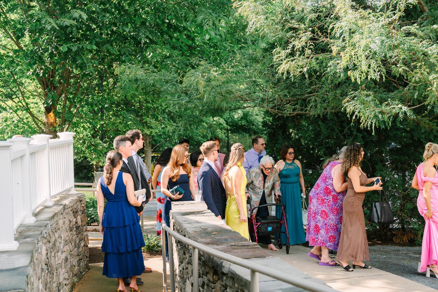 Colorfully dressed wedding guests socializing on a stone terrace surrounded by lush green trees.