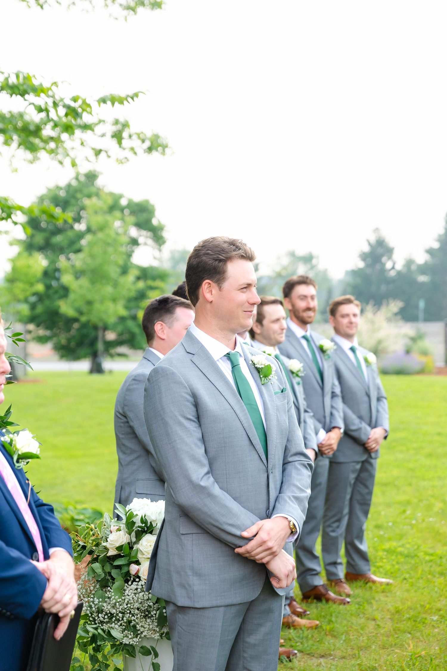Groom in gray suit with green tie smiling as bride walks down outdoor wedding ceremony aisle.