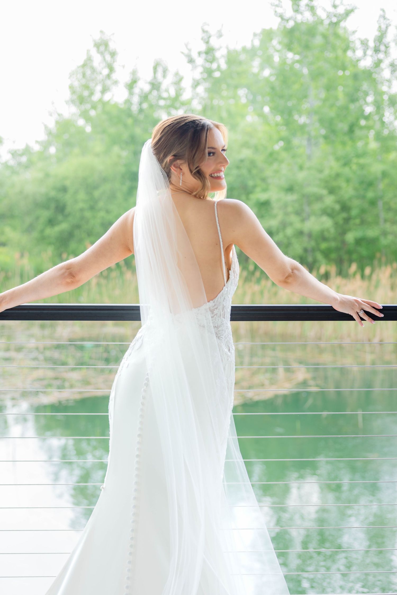 Smiling bride in backless lace wedding dress with veil poses on outdoor balcony near water.