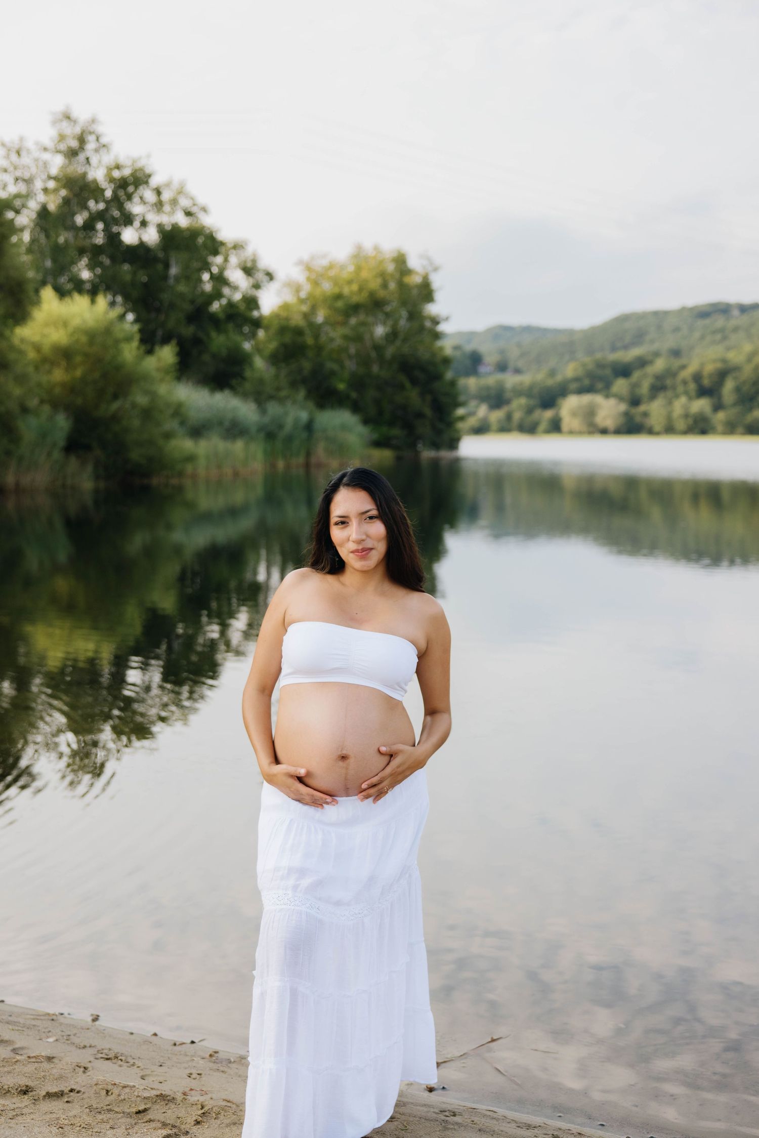 Pregnant woman in white tube top and skirt cradling her baby bump by a serene lake surrounded by lush greenery.