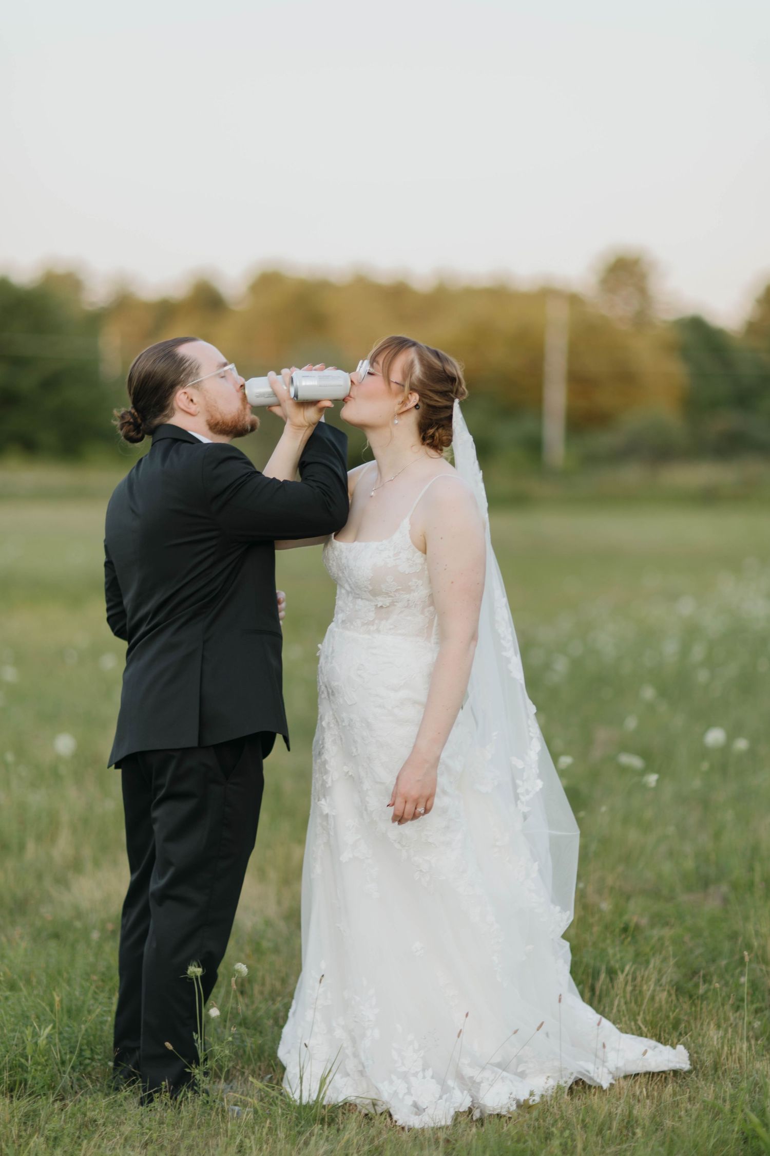 Bride and groom sharing a drink from white cans in a green field during golden hour wedding photos.