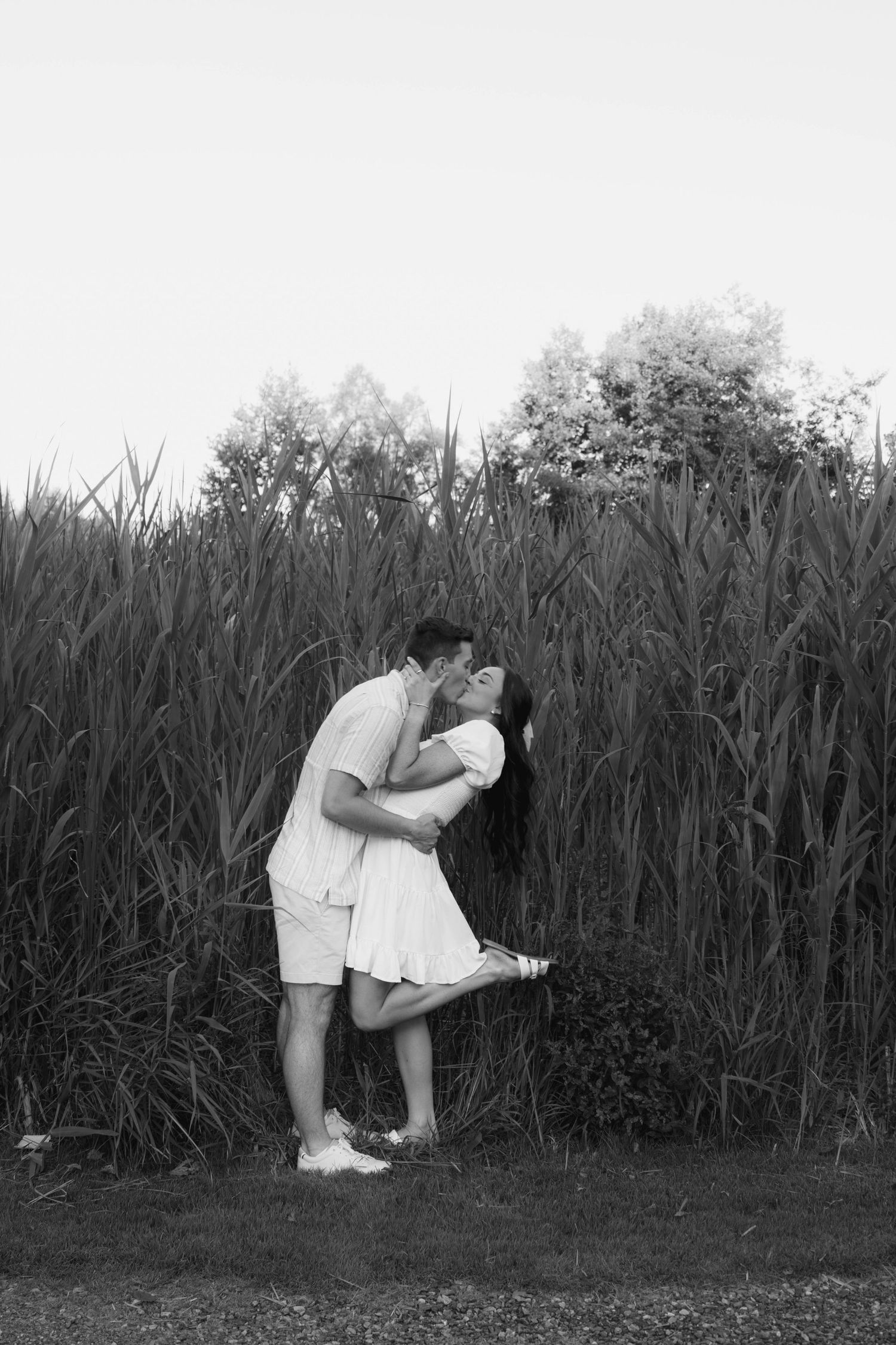 Black and white photo of couple sharing a romantic kiss near tall grass field.